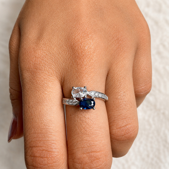 A close-up of a hand wearing a silver ring set with a clear round-cut center stone and a dark blue oval-cut center stone, with a matching silver band featuring small clear stones.