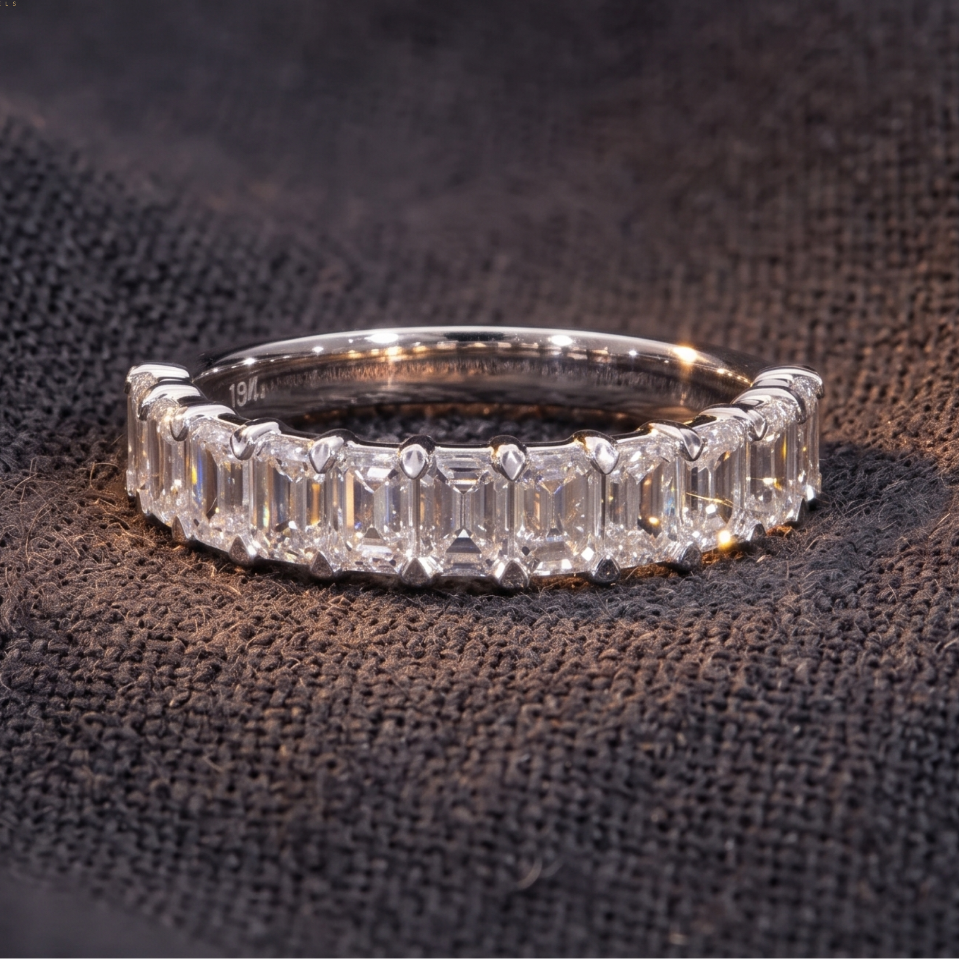 A silver band ring featuring a row of baguette-cut clear gemstones, photographed on a dark textured fabric background. The WITORIA logo is visible in the top-left corner.