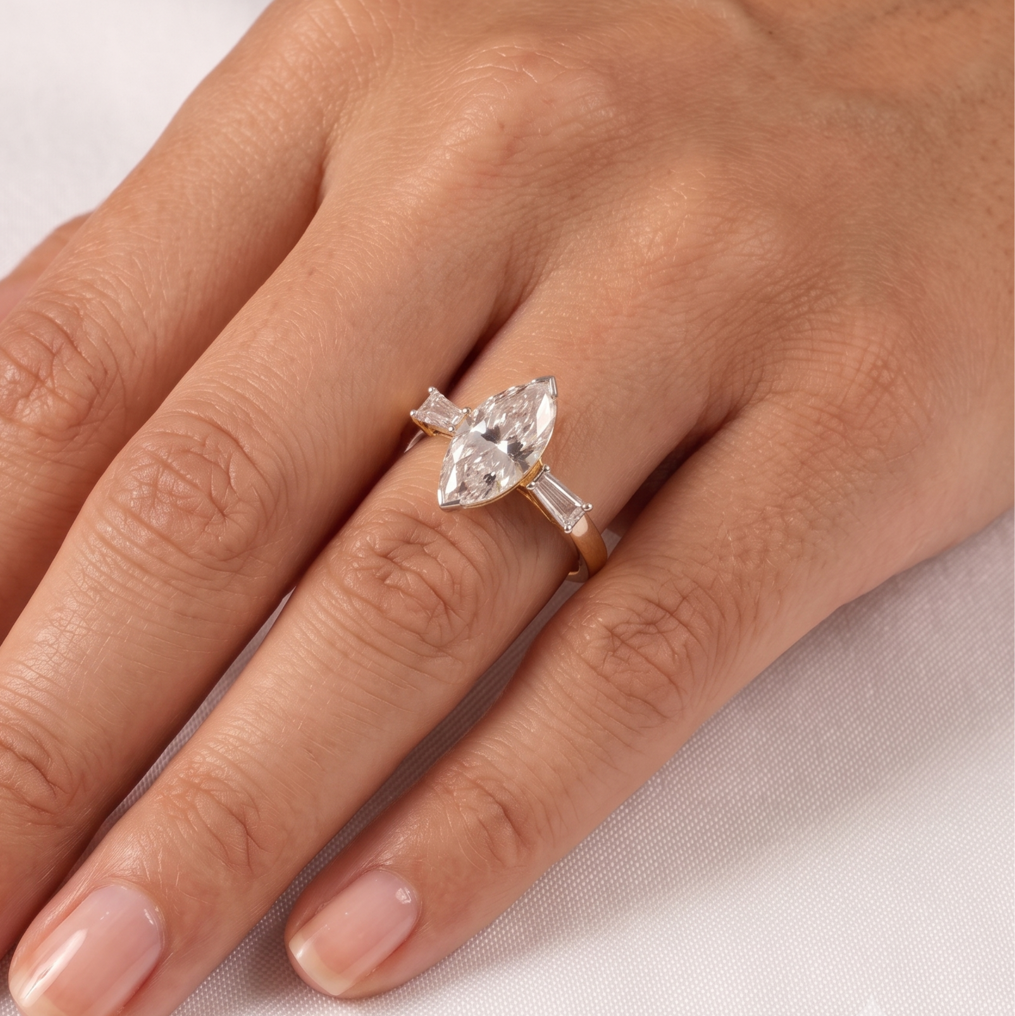 A close-up of a hand wearing a gold ring with a marquise-cut clear gemstone and baguette side stones on a light fabric background.