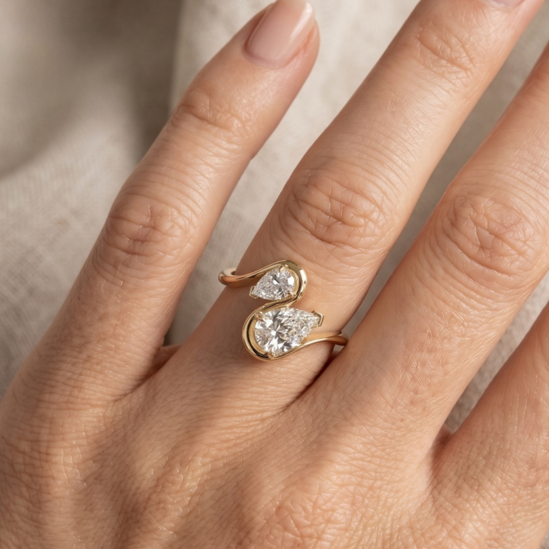 A close-up of a hand wearing a gold ring with two pear-cut clear gemstones in a swirled setting.