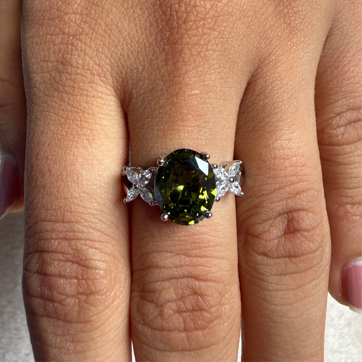 A close-up of a hand wearing a silver ring with a large oval green gemstone and small clear accent stones on either side of the band.