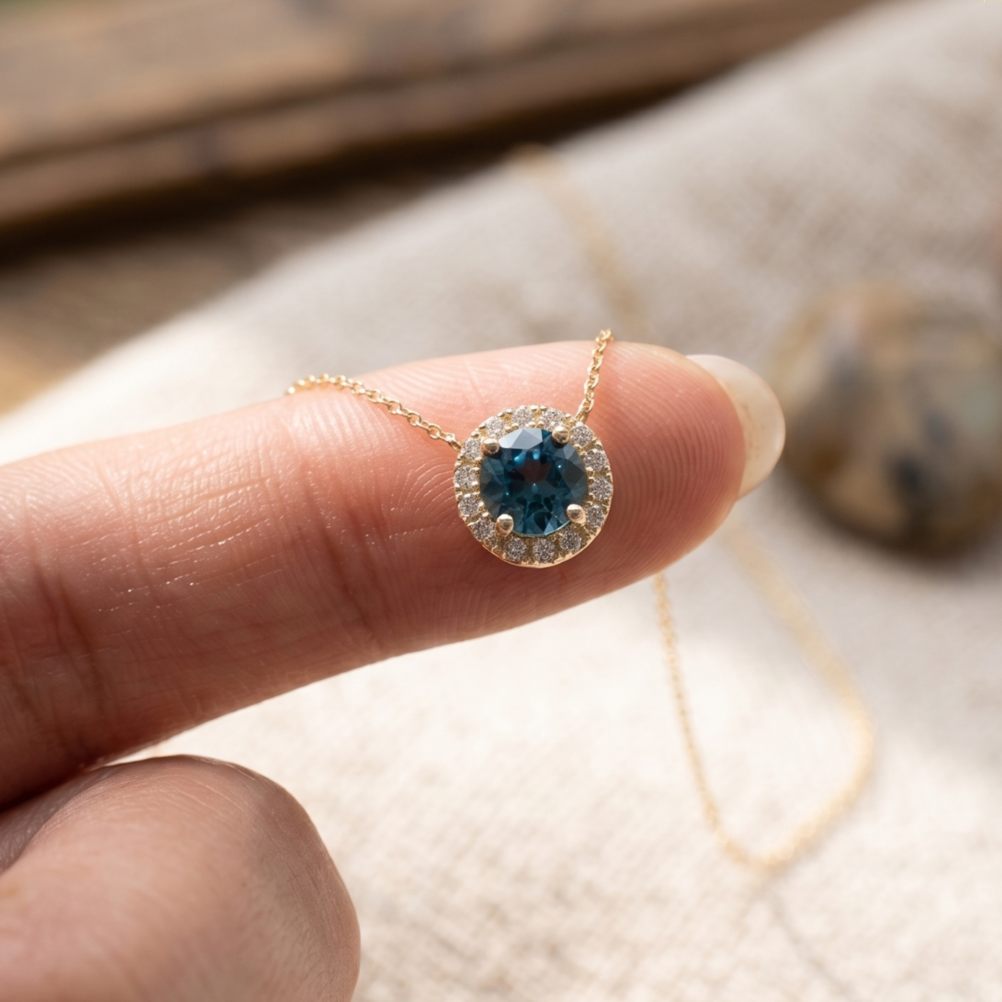 A gold necklace with a round blue gemstone surrounded by small clear stones, held on a person's finger against a soft neutral background.