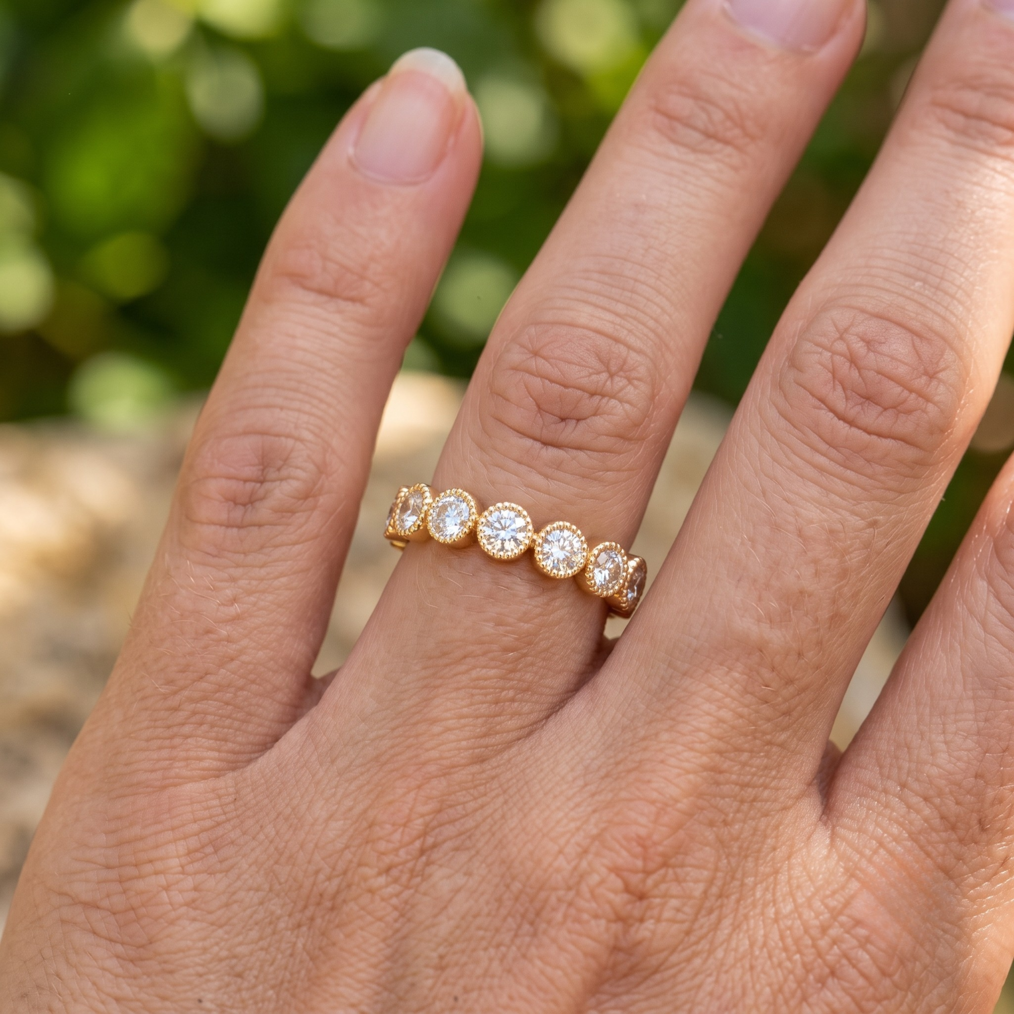 A close-up of a hand wearing a yellow gold eternity ring set with five round-cut clear gemstones, photographed outdoors with a blurred green background.
