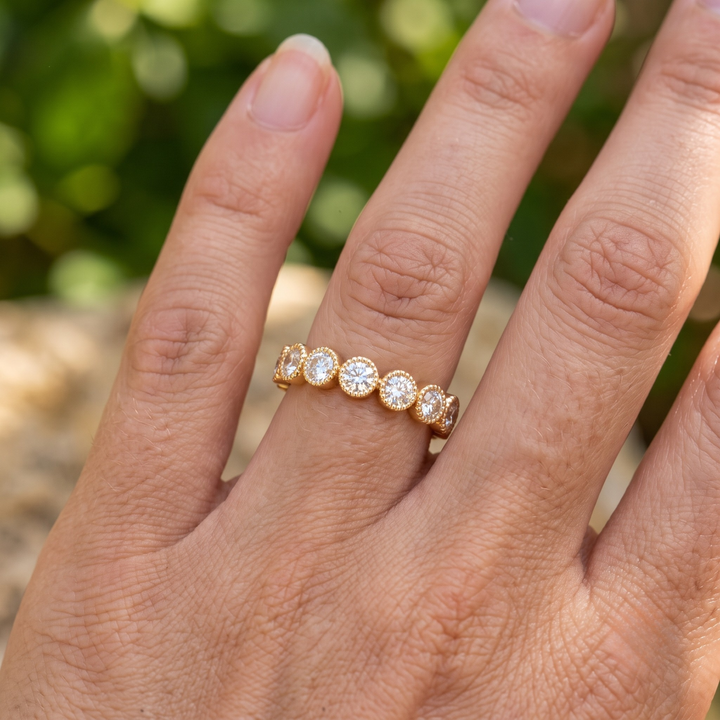 A close-up of a hand wearing a yellow gold eternity ring set with five round-cut clear gemstones, photographed outdoors with a blurred green background.