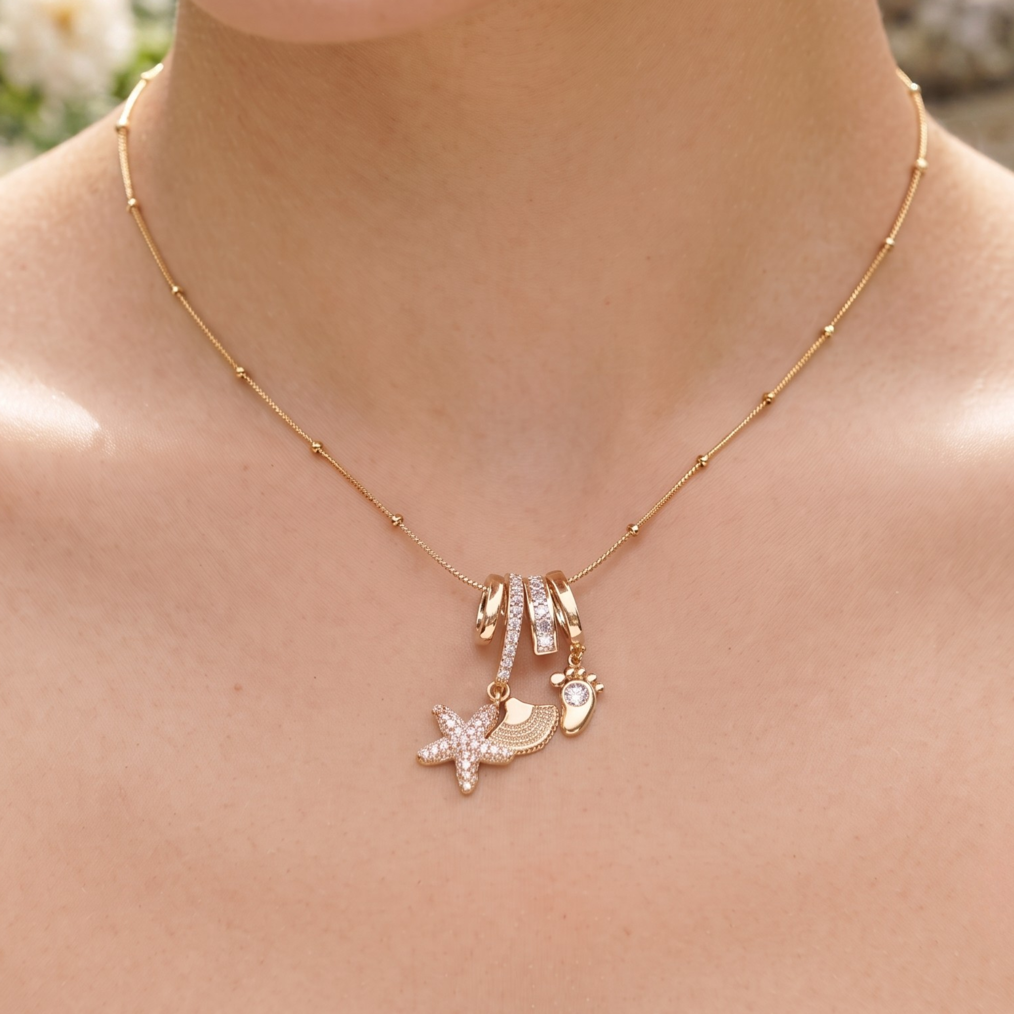 A close-up of a person's neck and chest wearing a delicate gold necklace with a starfish, seashell, and seashell-with-stone charm, accented by a small gold barrette with clear rhinestones.