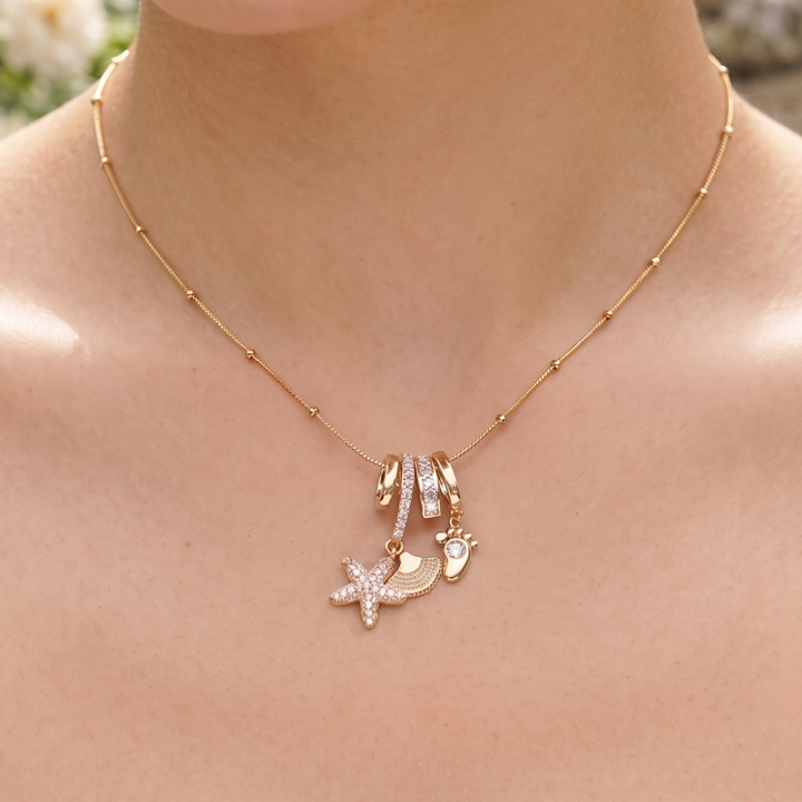 A close-up of a person's neck and chest wearing a delicate gold necklace with a starfish, seashell, and seashell-with-stone charm, accented by a small gold barrette with clear rhinestones.
