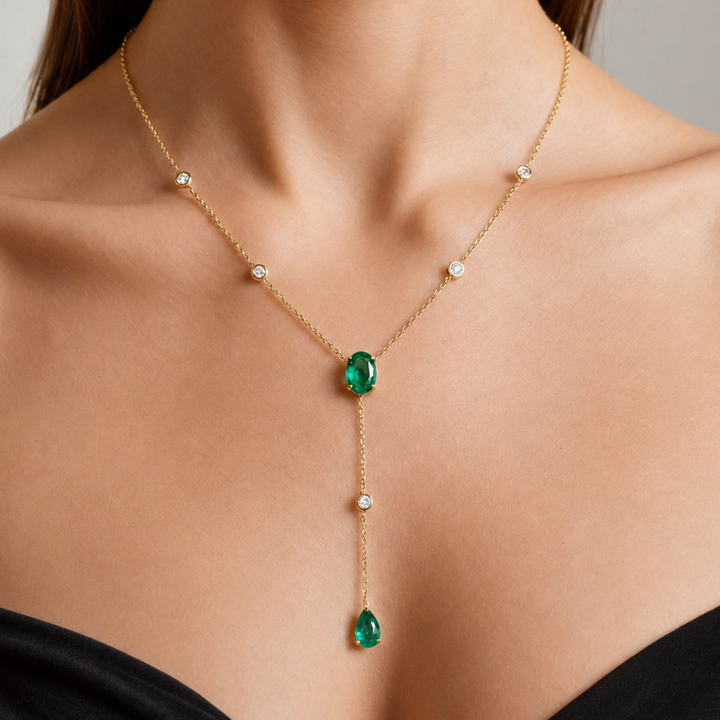 A gold Y-necklace with a large oval green emerald center stone and a dangling pear-shaped green emerald, worn on a model's neck.