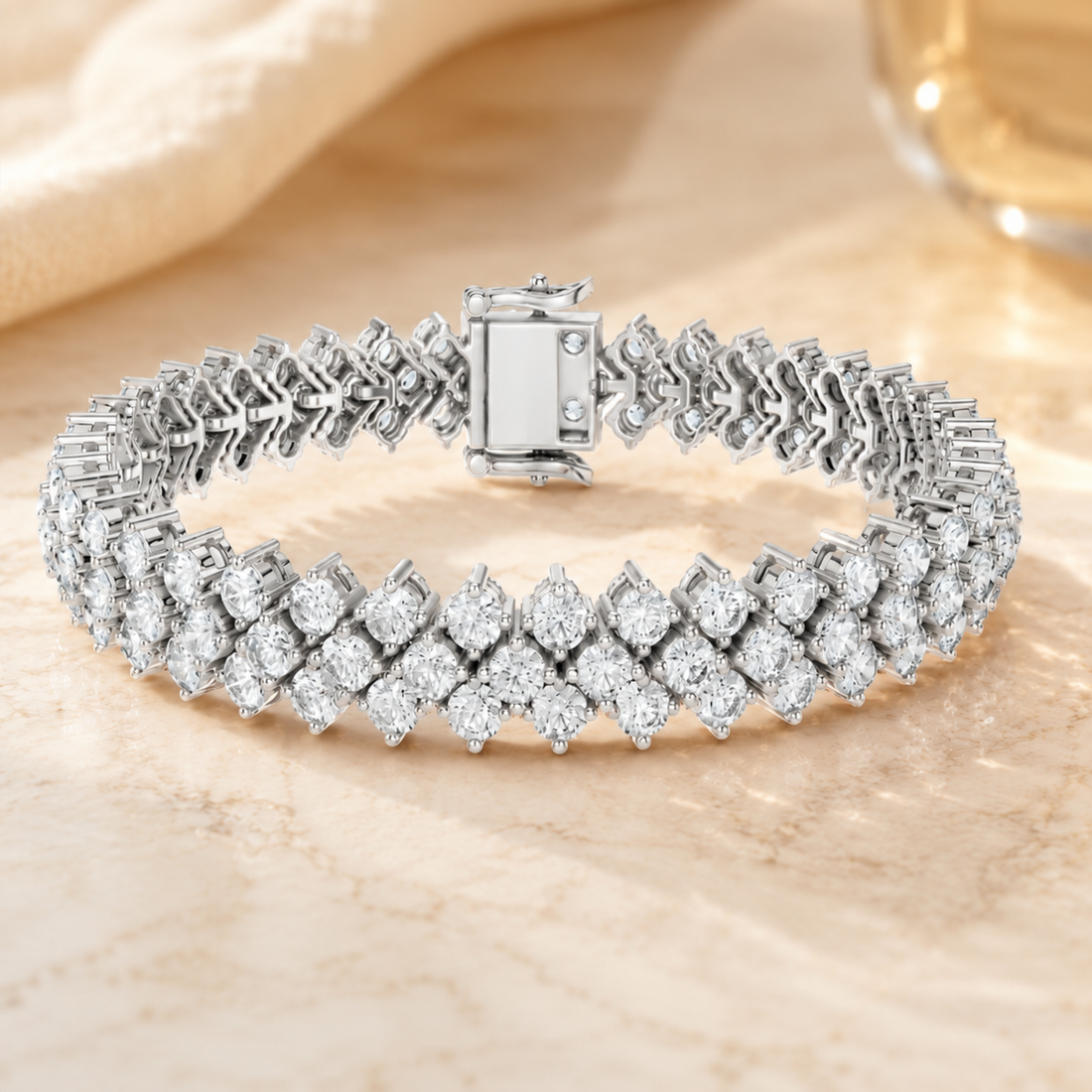 A diamond tennis bracelet in a silver-toned metal setting, displayed on a light beige marble surface with a soft beige fabric and glass in the background.