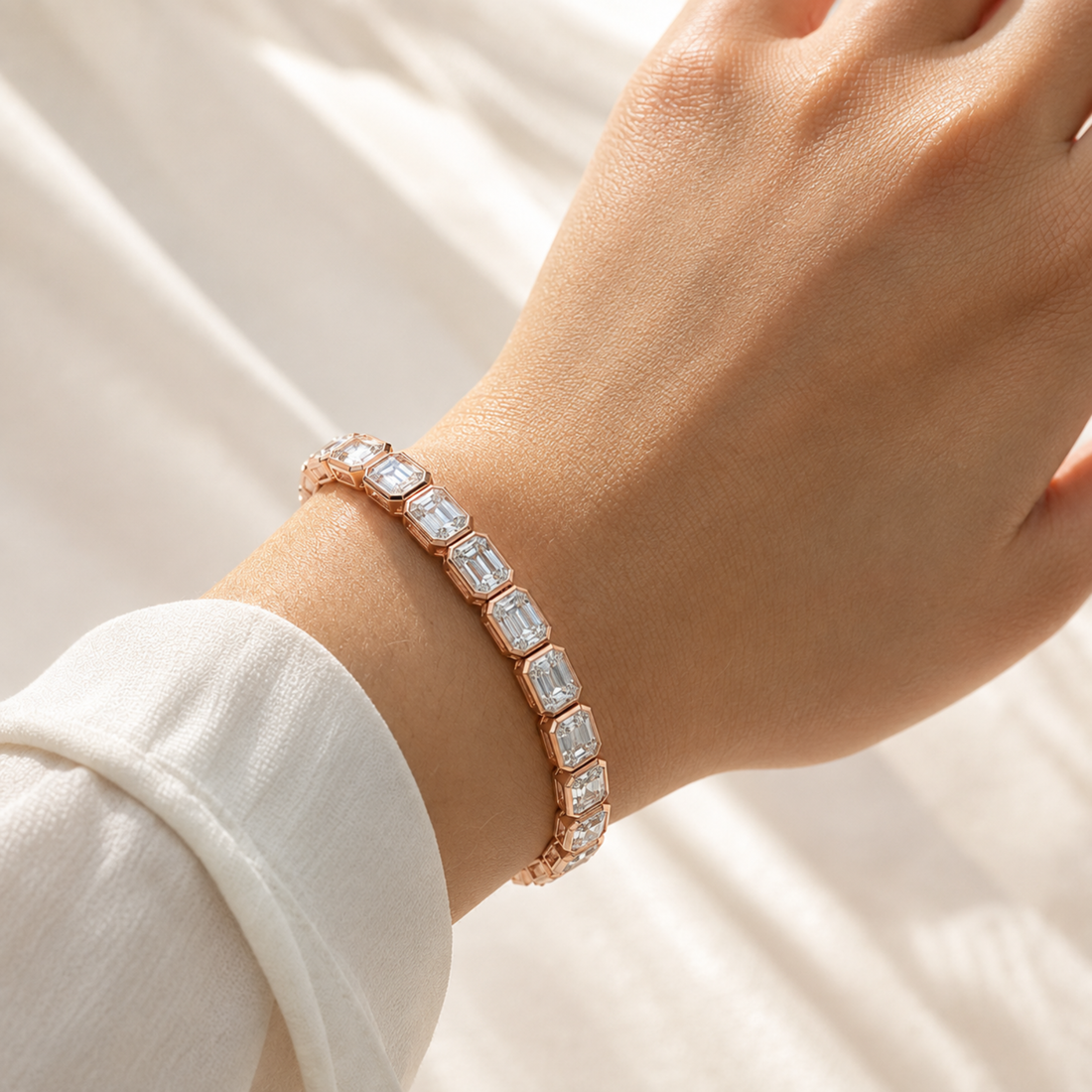 A close-up of a person's wrist wearing a rose gold bracelet with Emerald cut clear crystals, set against a soft white fabric background.