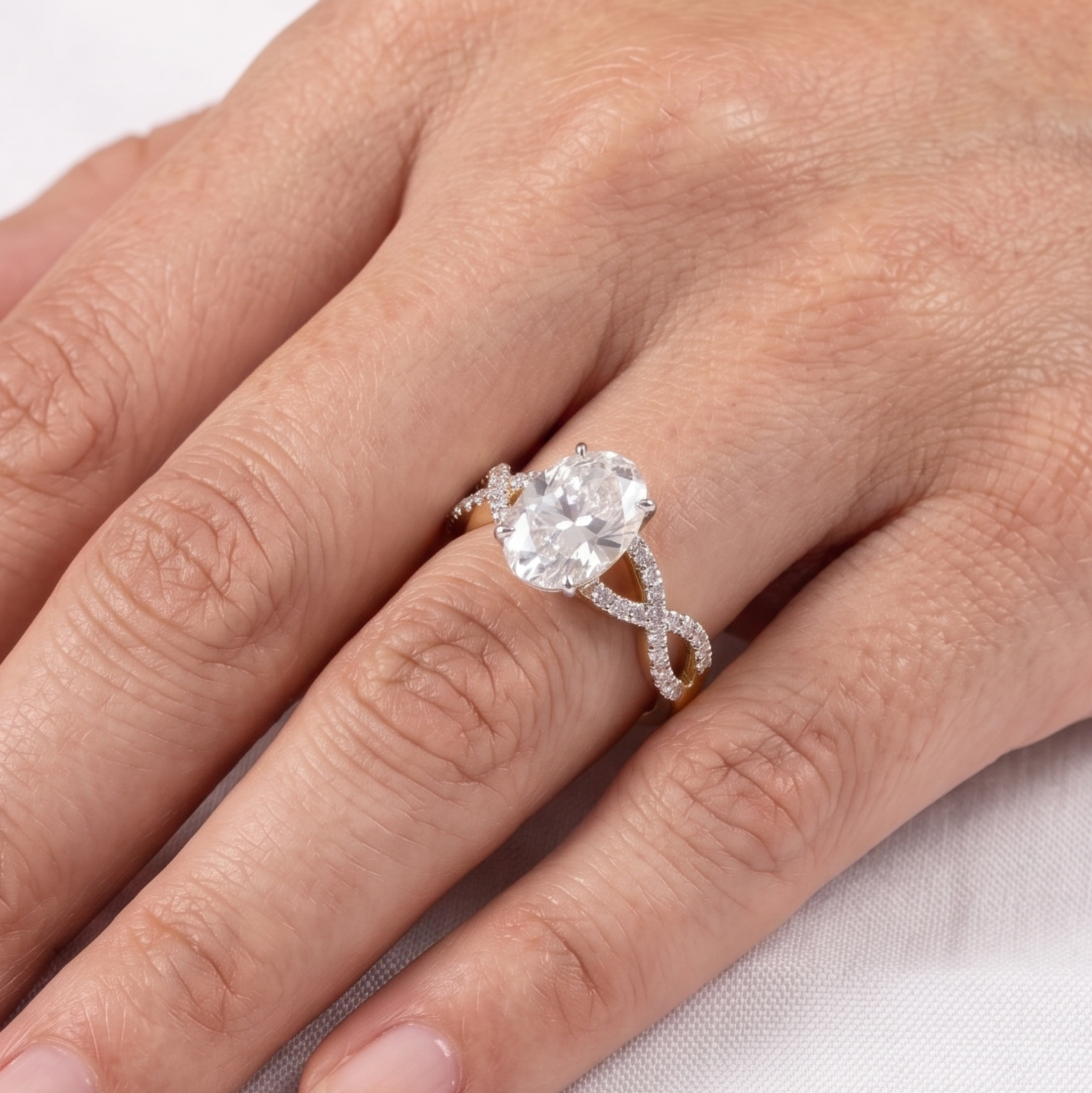 A yellow gold engagement ring featuring a large oval-cut clear center stone and pavé-set accent stones on the band, photographed on a textured white fabric background.