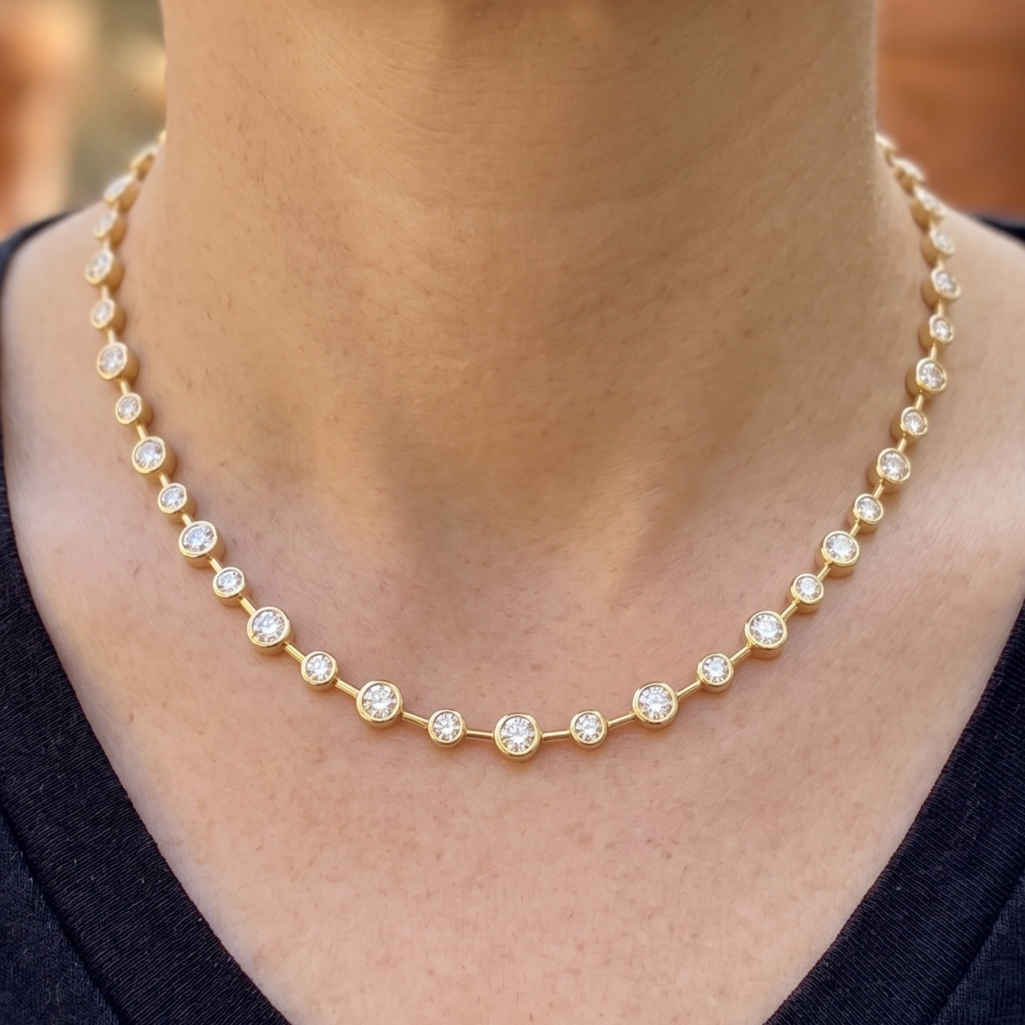 A close-up of a person wearing a delicate gold tennis necklace with small clear round-cut stones set in each link. The necklace is shown against the neck and upper chest, with a dark navy or black top visible at the bottom.