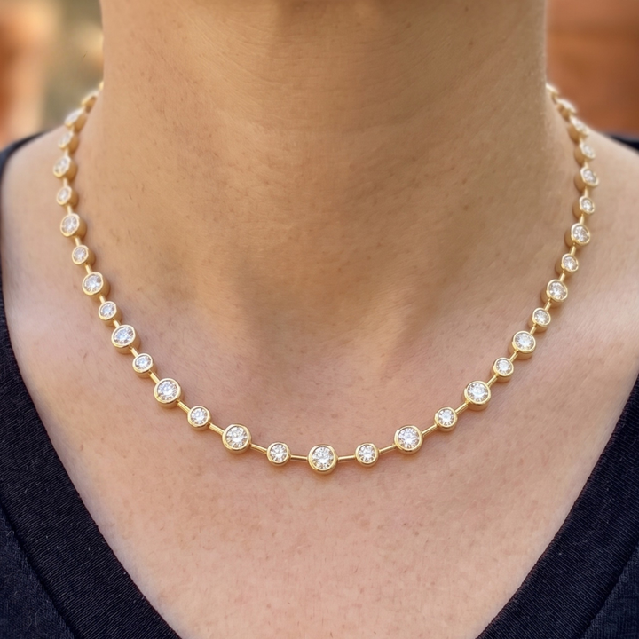 A close-up of a person wearing a delicate gold tennis necklace with small clear round-cut stones set in each link. The necklace is shown against the neck and upper chest, with a dark navy or black top visible at the bottom.