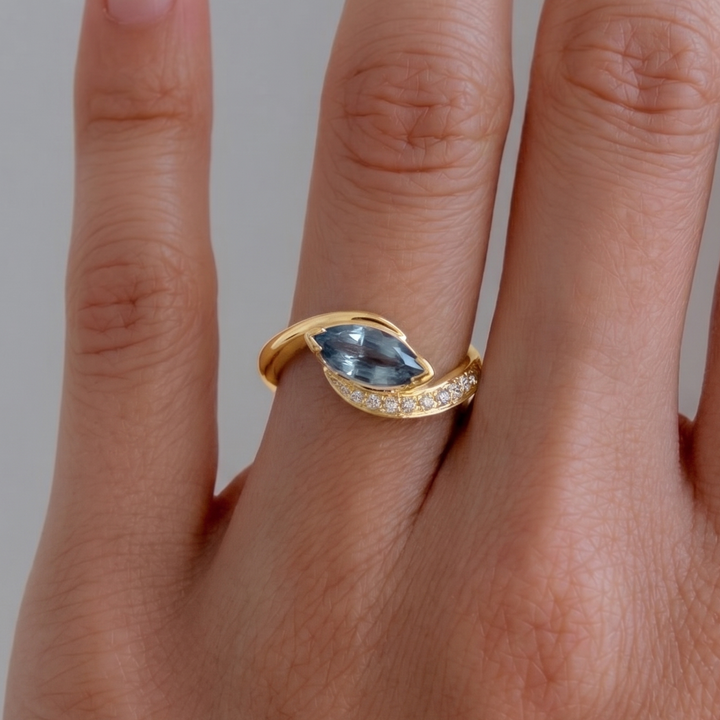 A gold ring with a marquise-cut light blue gemstone and a curved row of small clear stones, shown worn on a person's finger against a soft gray background.