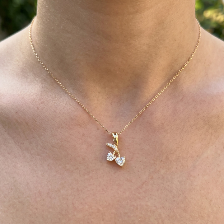 A delicate gold chain necklace with a small bow-shaped pendant set with clear stones, shown worn on a person's neck against a softly blurred outdoor background.