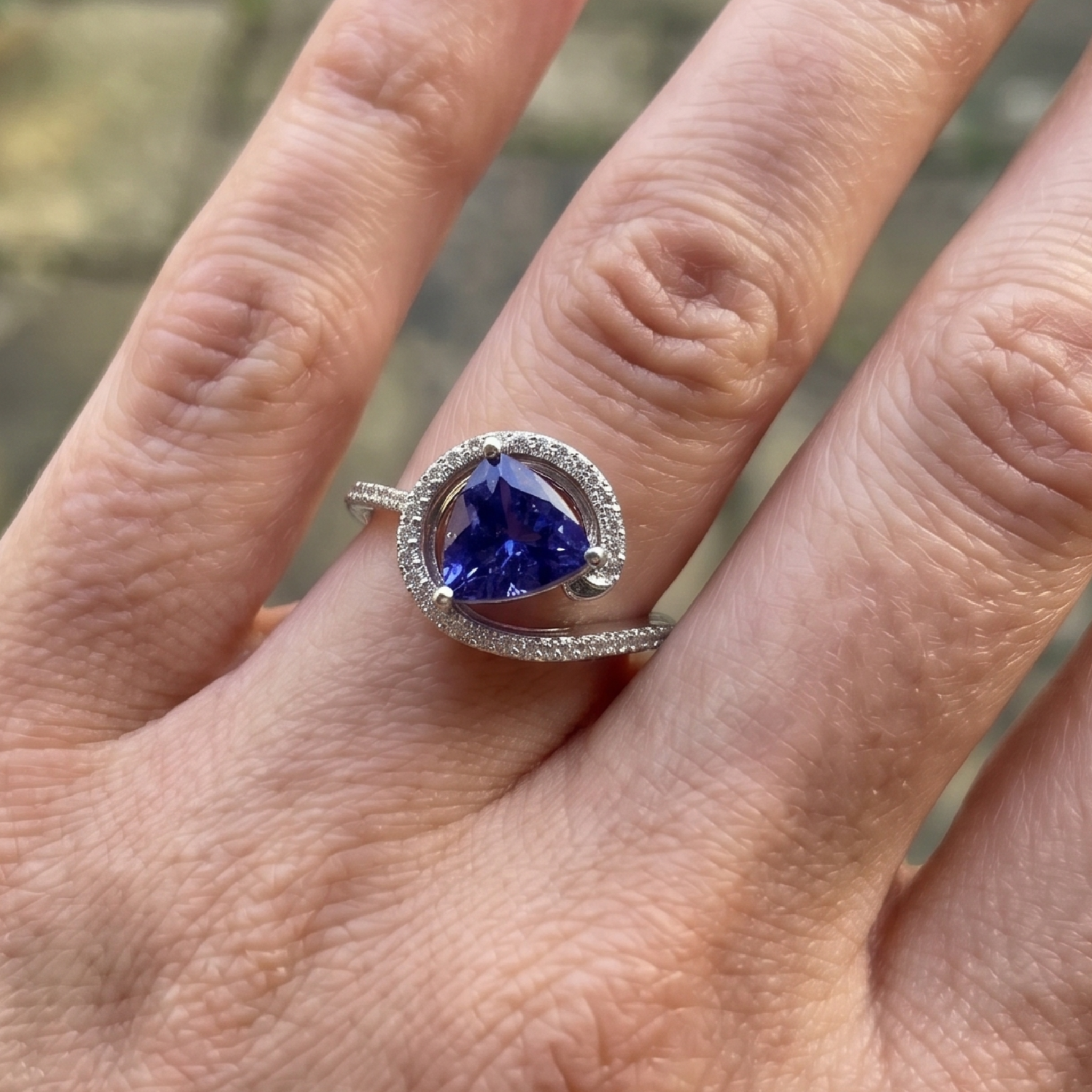 A silver ring featuring a triangular deep blue gemstone surrounded by a halo of small clear stones, worn on a person's finger.
