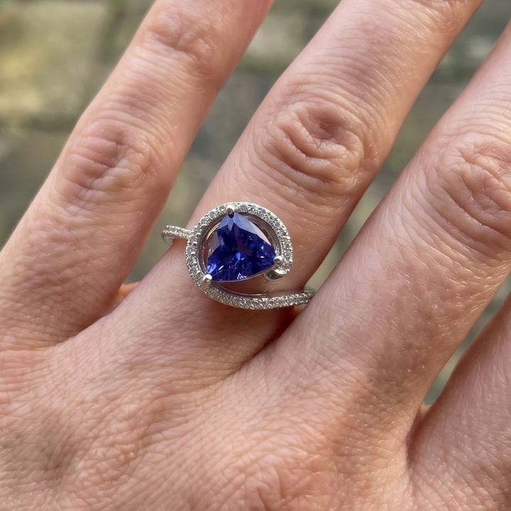 A silver ring featuring a triangular deep blue gemstone surrounded by a halo of small clear stones, worn on a person's finger.