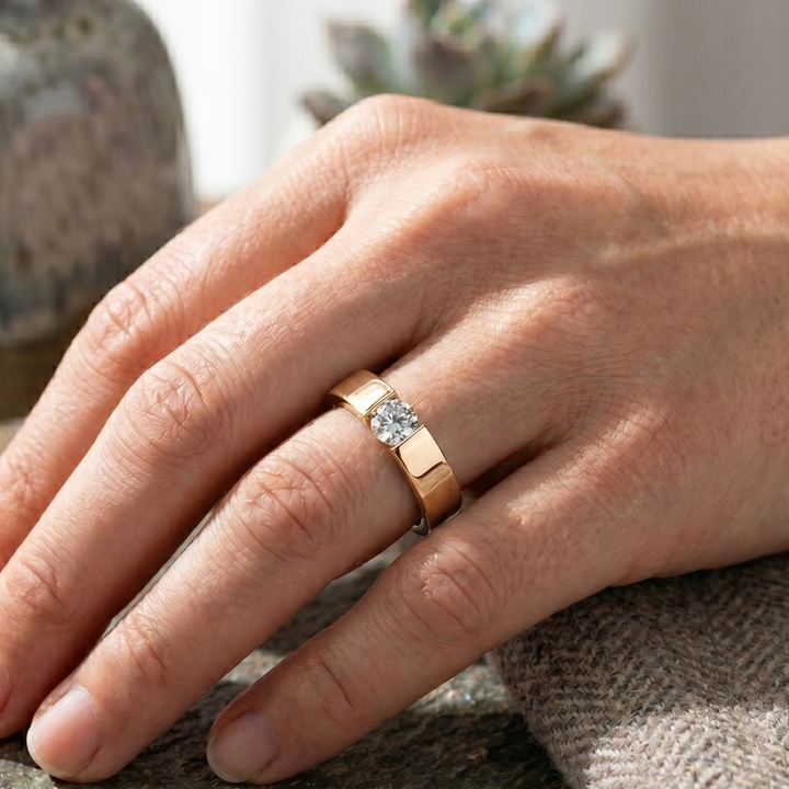 A close-up of a hand wearing a gold band ring with a square-cut clear gemstone in a prong setting, resting on a textured gray surface with a blurred background.
