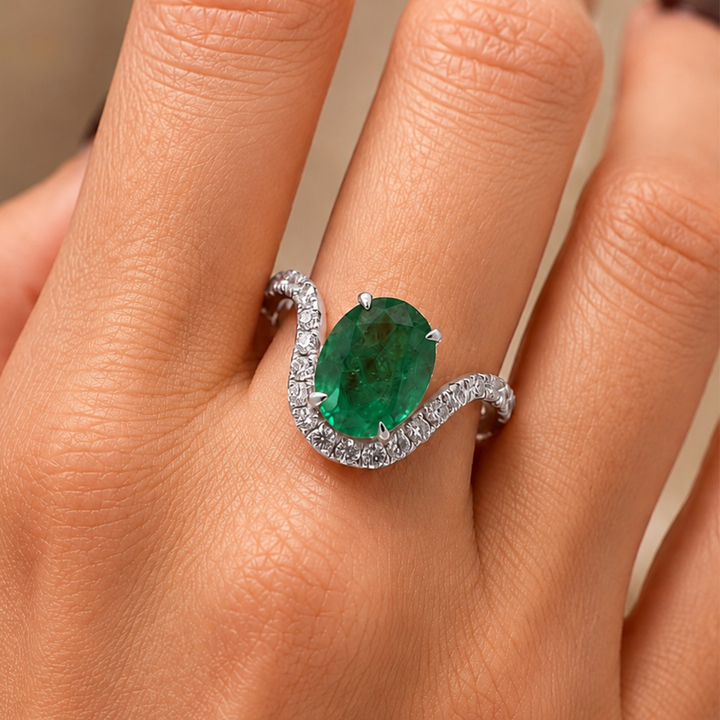 A close-up of a hand wearing a silver ring with a large oval green emerald center stone and a halo of small clear stones along a wavy band.