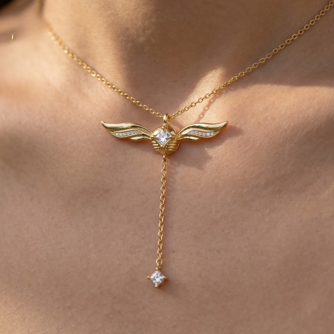 A gold winged pendant necklace with a dangling charm, worn on a model's neck. The pendant features a central clear gemstone and small clear stones on the wings, set against warm, sunlit skin.