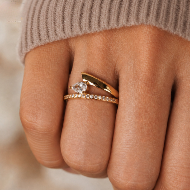 A close-up of a hand wearing two gold rings: one with a pear-cut clear gemstone and the other a delicate pavé band, styled with a ribbed taupe knit cuff.