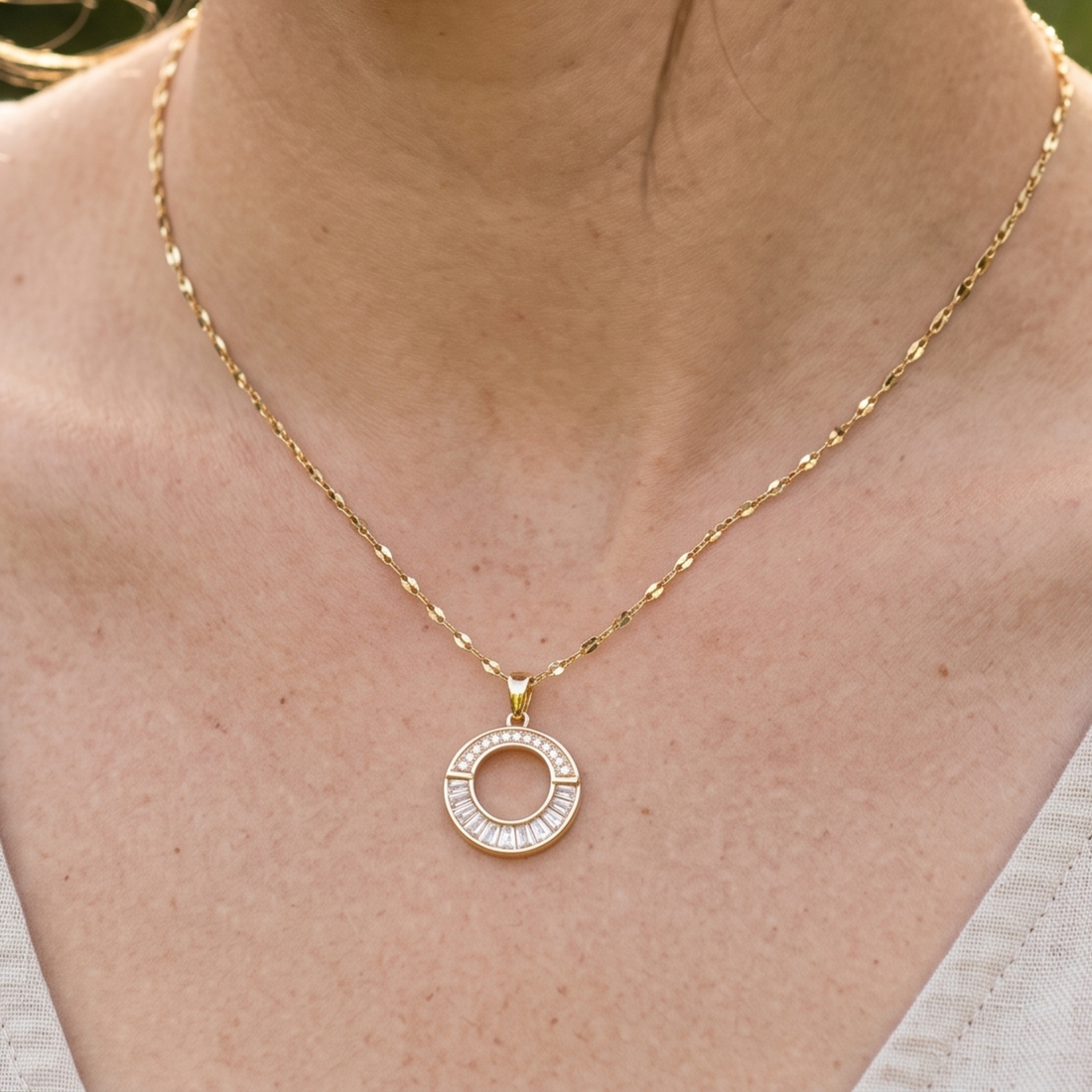 A gold necklace with a circular pendant featuring small clear stones, worn on a person's neck over a white top.