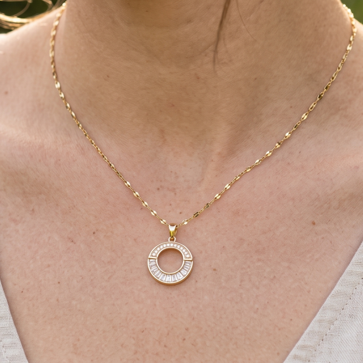 A gold necklace with a circular pendant featuring small clear stones, worn on a person's neck over a white top.