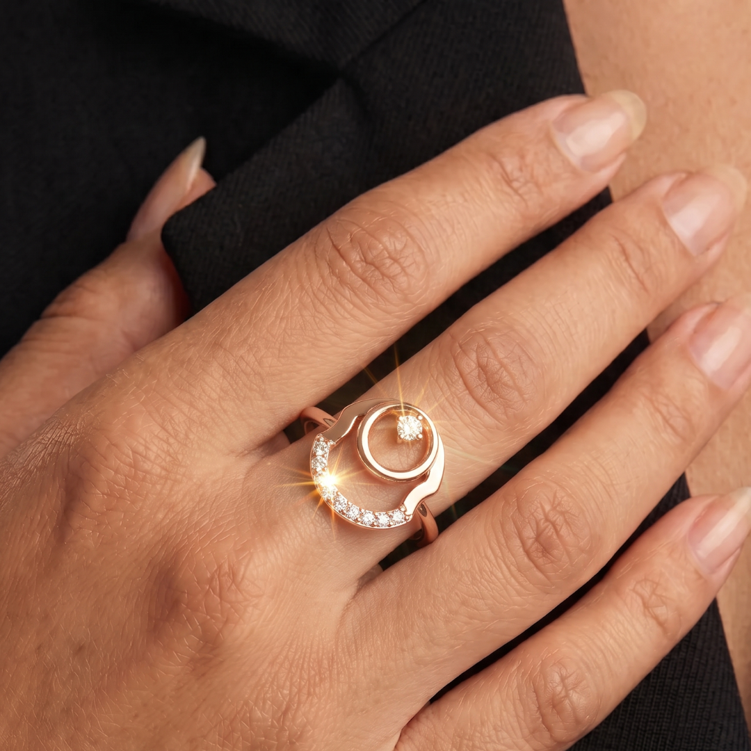 A close-up of a hand wearing a rose gold ring with a crescent moon design, a central round gemstone, and a smaller diamond-like accent on a black fabric background.