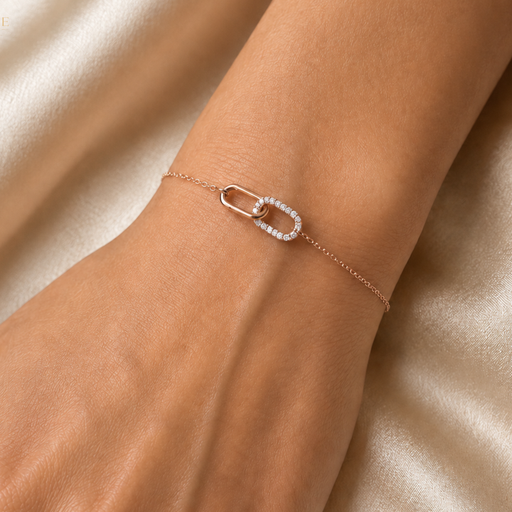 A delicate rose gold chain bracelet with a link design, one of which is paved with small clear stones, shown worn on a wrist against a soft beige satin background.