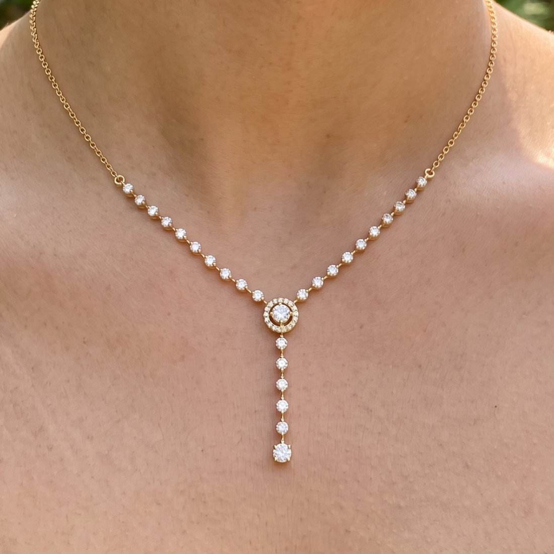 A delicate gold necklace with small clear stones along the chain and a central round stone, with a drop pendant, shown worn on a person's neck and upper chest.