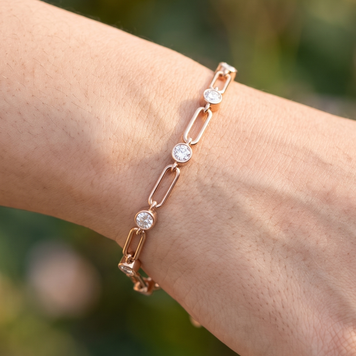 A rose gold chain bracelet with three round-cut clear gemstones, shown worn on a person's wrist against a soft, blurred green background.