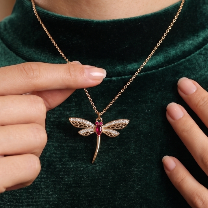 A close-up of a gold dragonfly pendant necklace with a red gemstone, worn by a person in a dark green velvet top. The person's hands are visible holding the chain and framing the necklace.