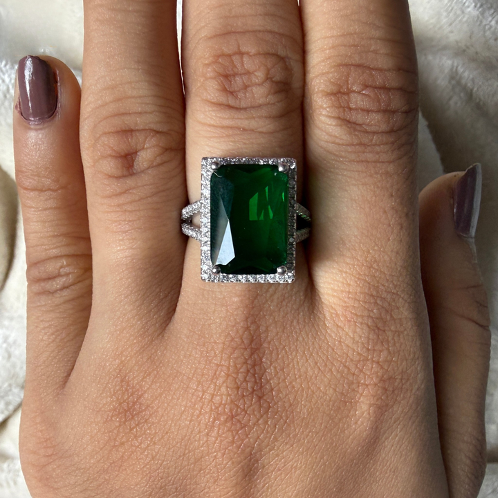 A rectangular emerald-cut green gemstone ring with a halo of small clear stones, shown worn on a person's finger.