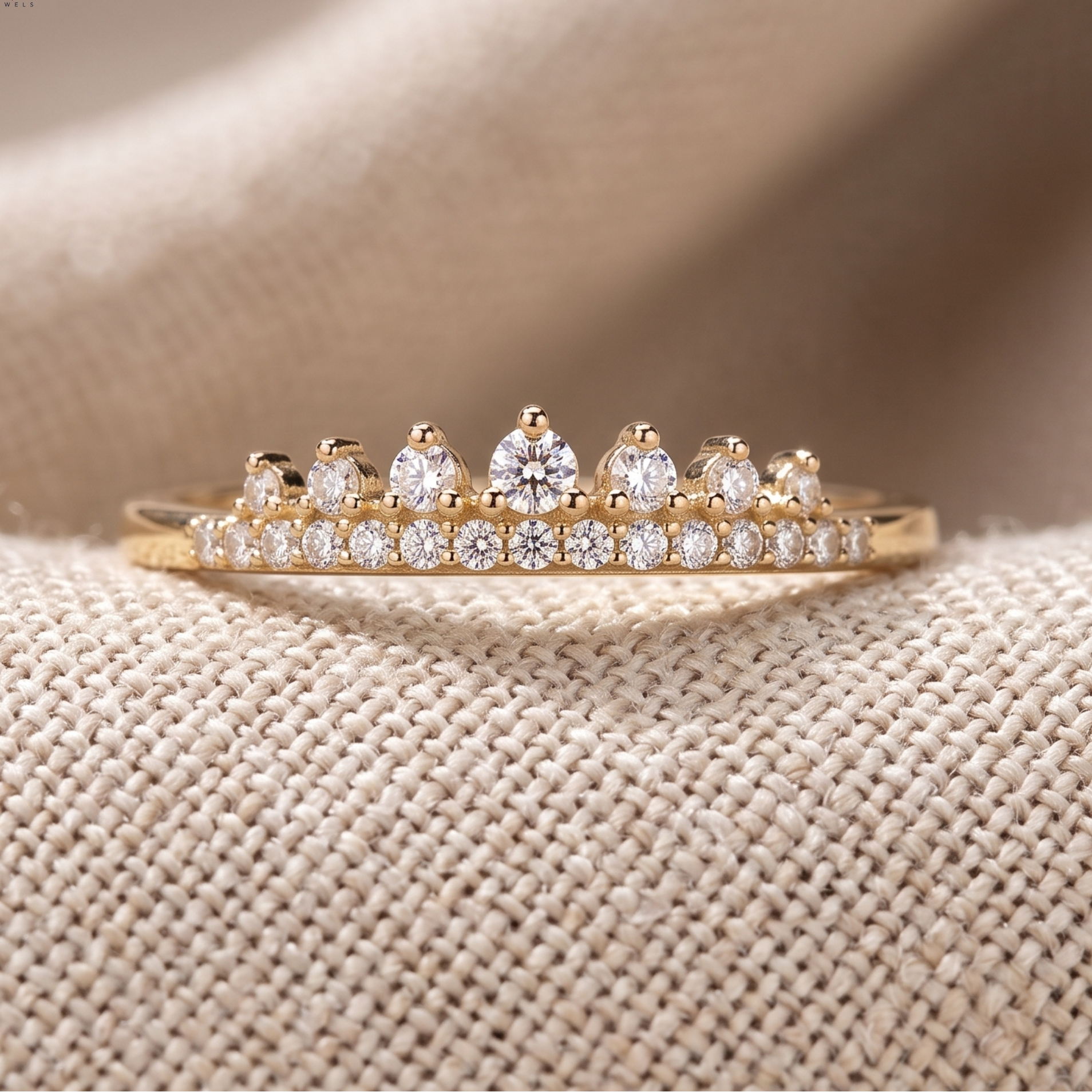 A gold ring featuring a row of clear round diamonds and five pear-cut diamonds, displayed on a textured beige fabric background. The brand logo 'Widoria' is visible in the top left corner.