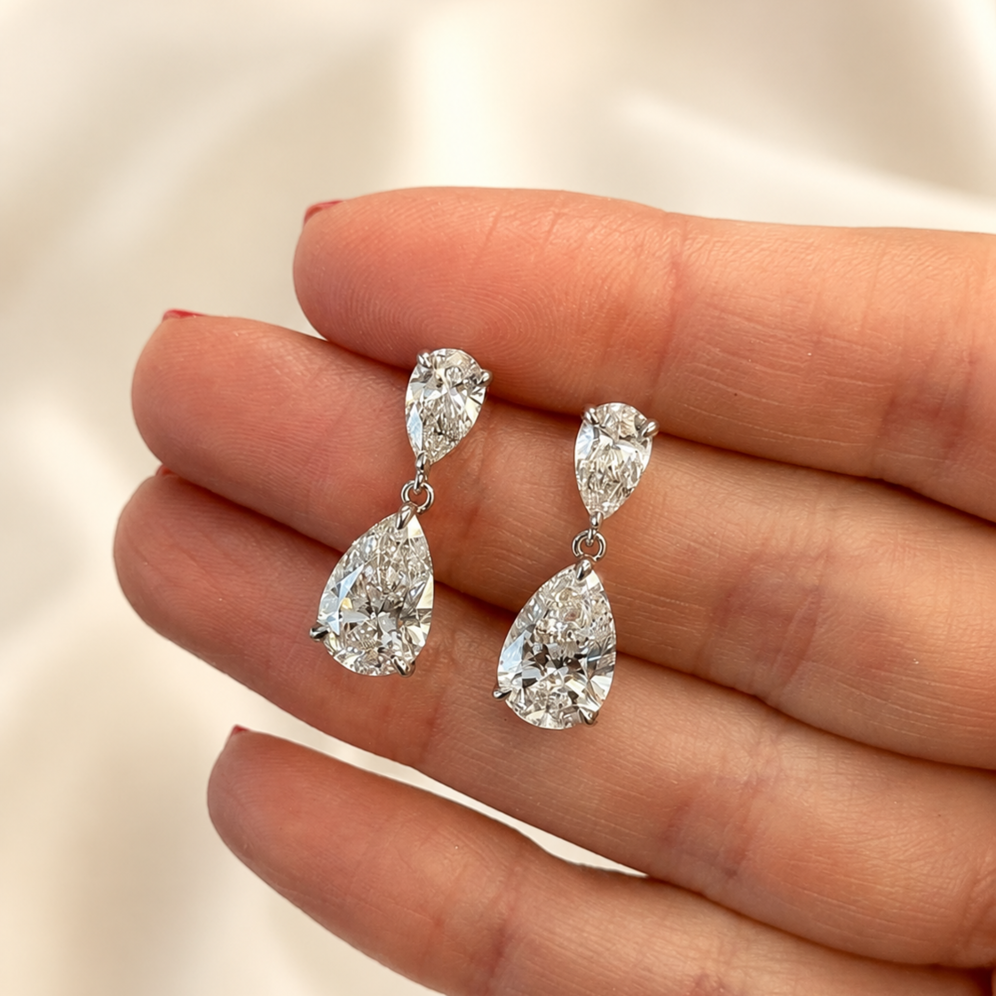A pair of silver-tone drop earrings with pear-cut clear gemstones, held on a person's fingers against a soft light background.