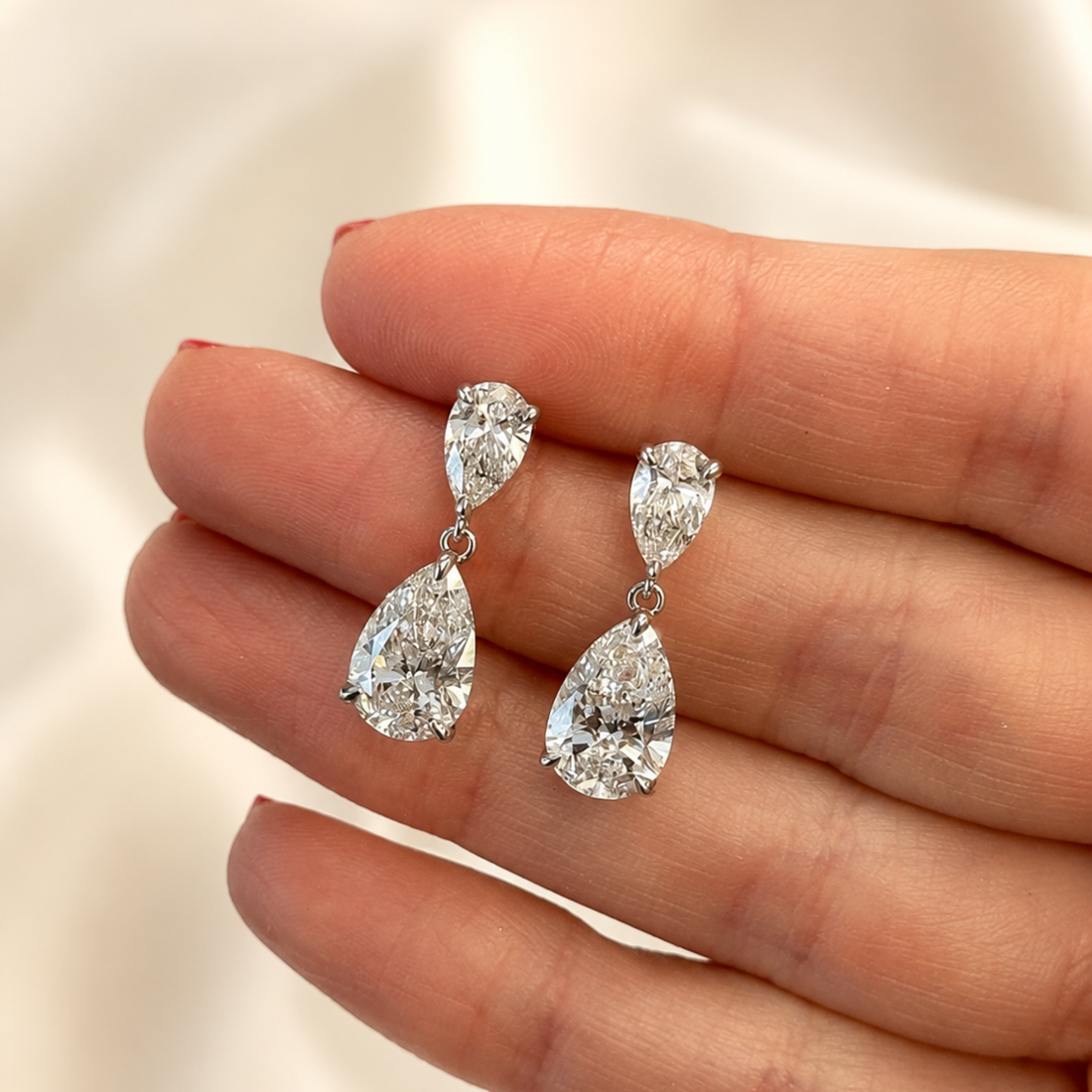 A pair of silver-tone drop earrings with pear-cut clear gemstones, held on a person's fingers against a soft light background.