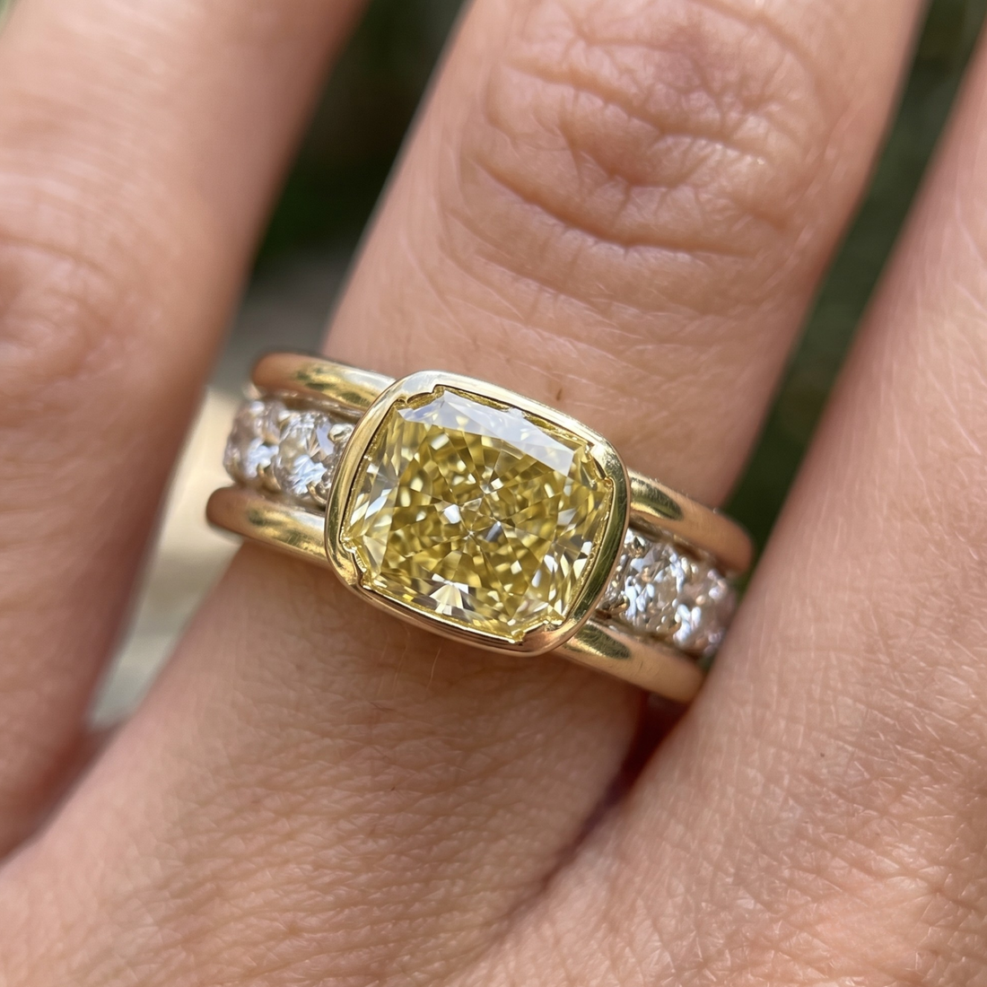 A yellow cushion-cut diamond ring with a yellow gold band and small clear pavé diamonds, shown worn on a finger.