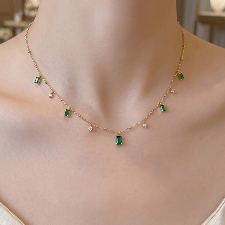 A delicate gold chain necklace with small green rectangular gemstones and tiny white pearl-like accents, shown worn on a model's neck.