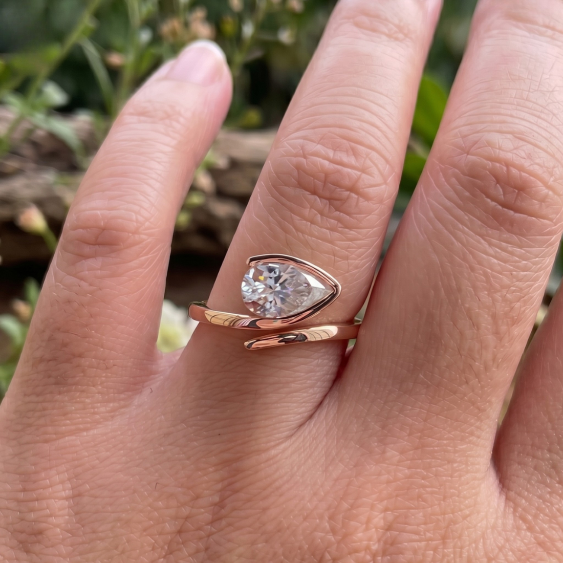 A close-up of a hand wearing a rose gold ring with a pear-cut clear gemstone in a bezel setting, with a wrap-around band.