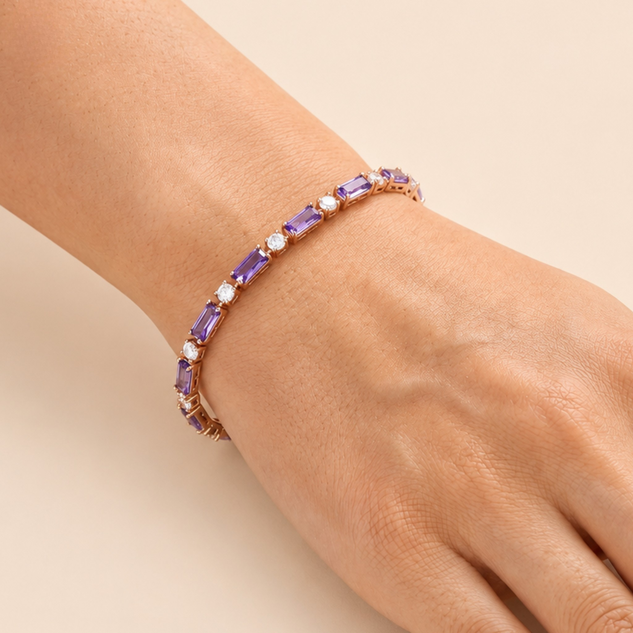 A close-up of a person's wrist wearing a delicate bracelet with purple rectangular gemstones and small white stones set in a rose gold-toned band, against a soft beige background.