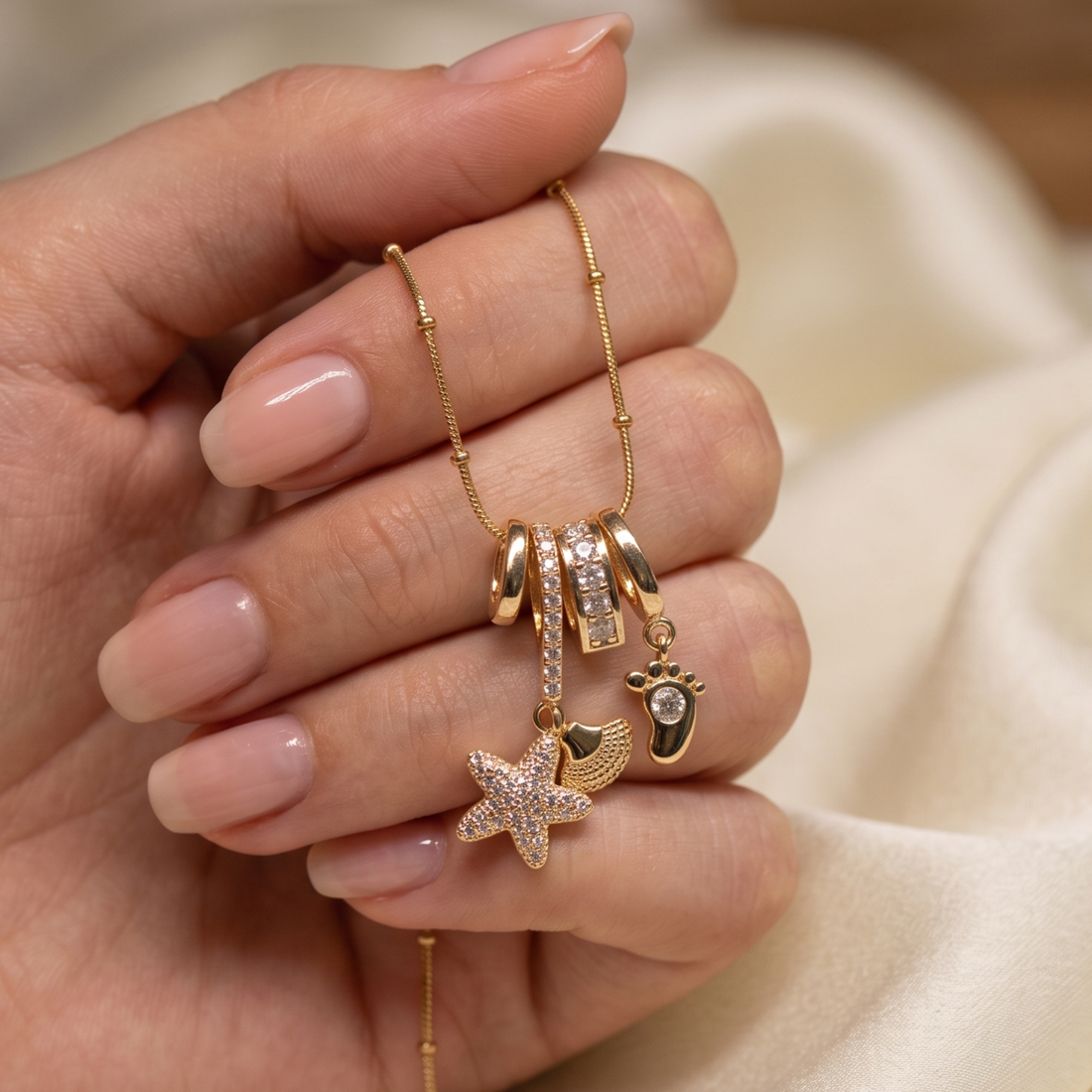 A close-up of a hand holding a delicate gold chain necklace with multiple charms, including a starfish, a crescent moon, a small heart, and a baby bootie charm, against a soft cream-colored fabric background.