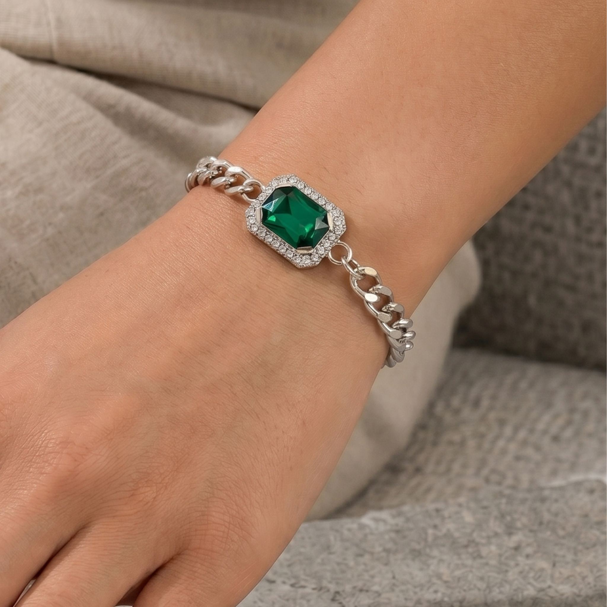 A close-up of a person's wrist wearing a silver-toned chain bracelet with a central square green gemstone surrounded by small clear stones.