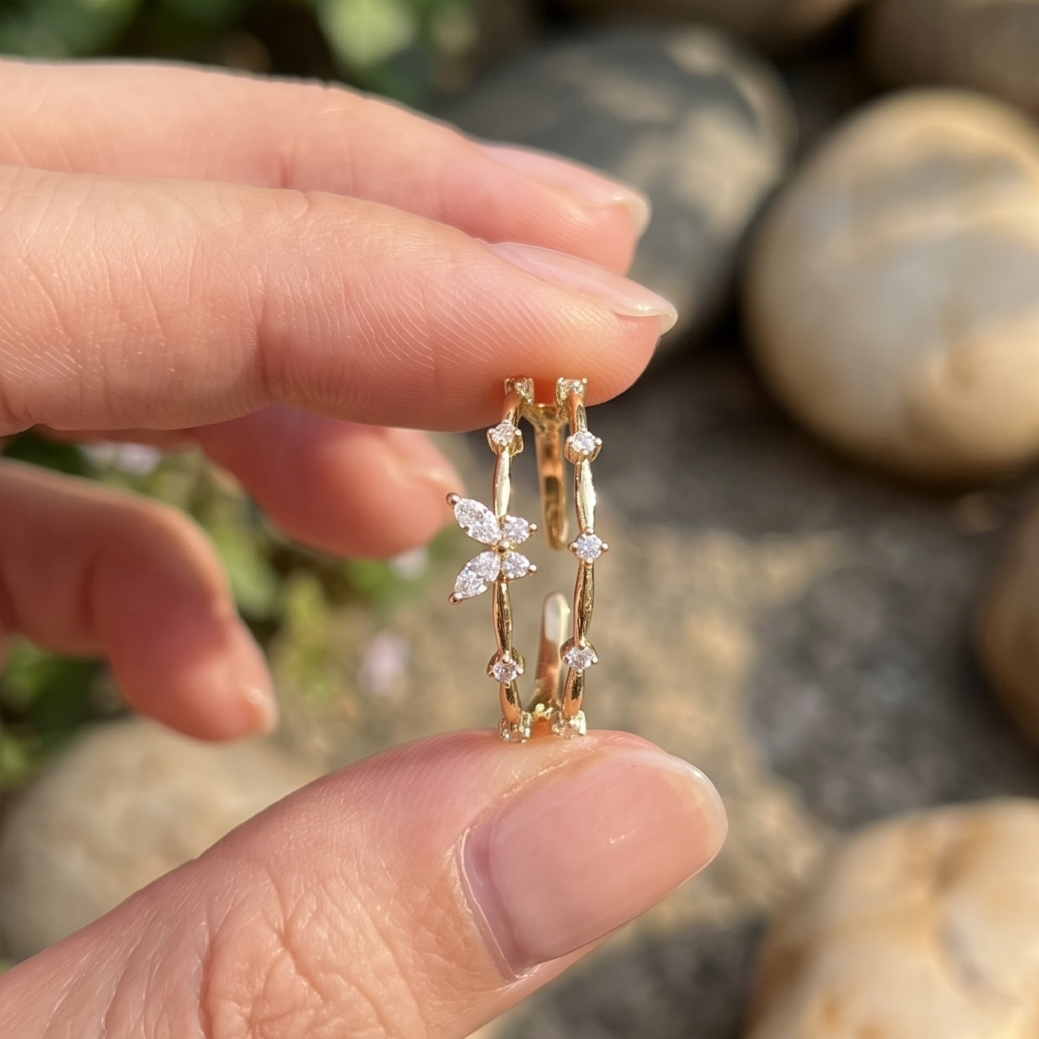 A close-up of a hand holding a delicate gold ring with a butterfly and diamond design, set against a blurred outdoor background of rocks.
