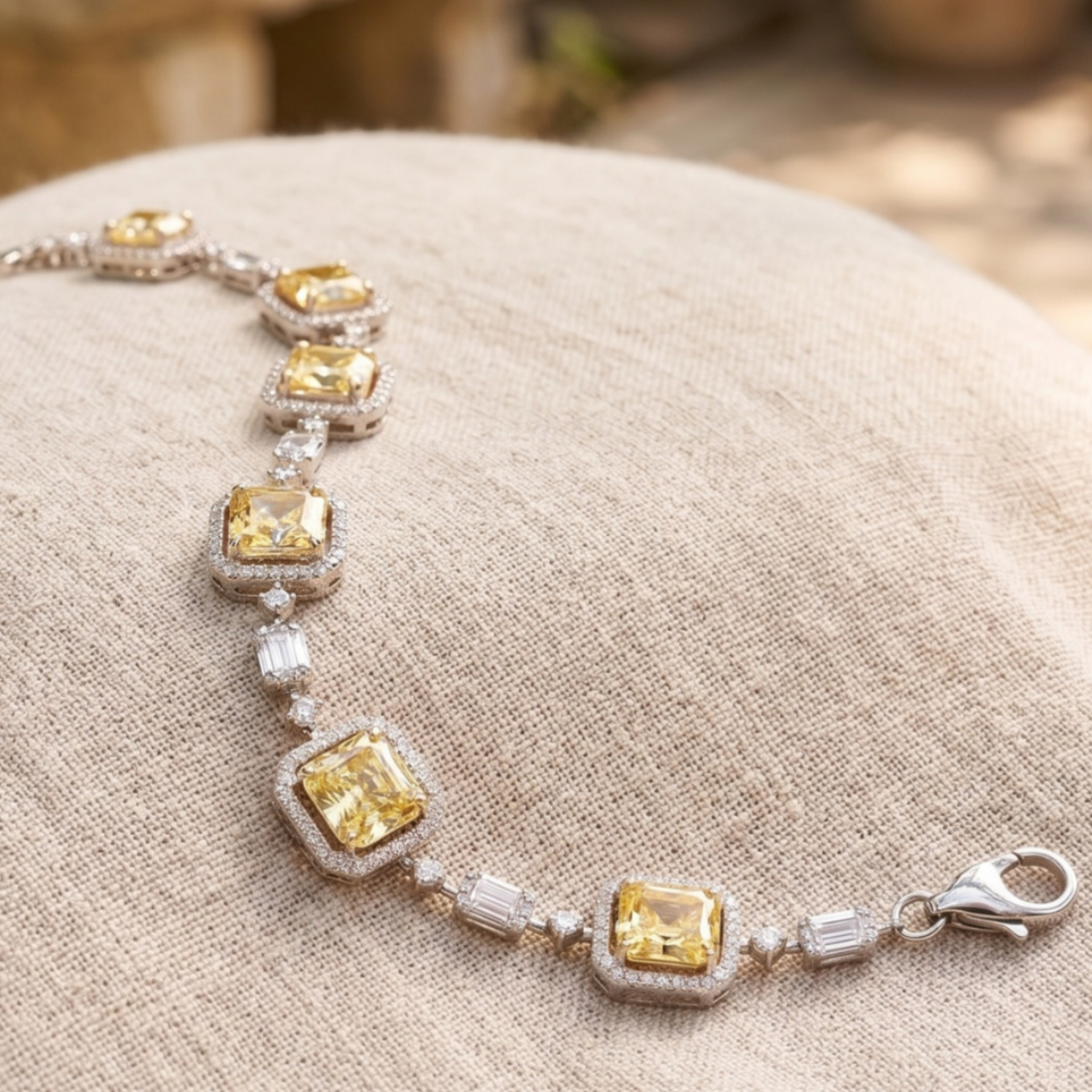 A silver-toned bracelet featuring five large yellow cushion-cut center stones, each surrounded by small clear stones, displayed on a textured beige fabric surface.