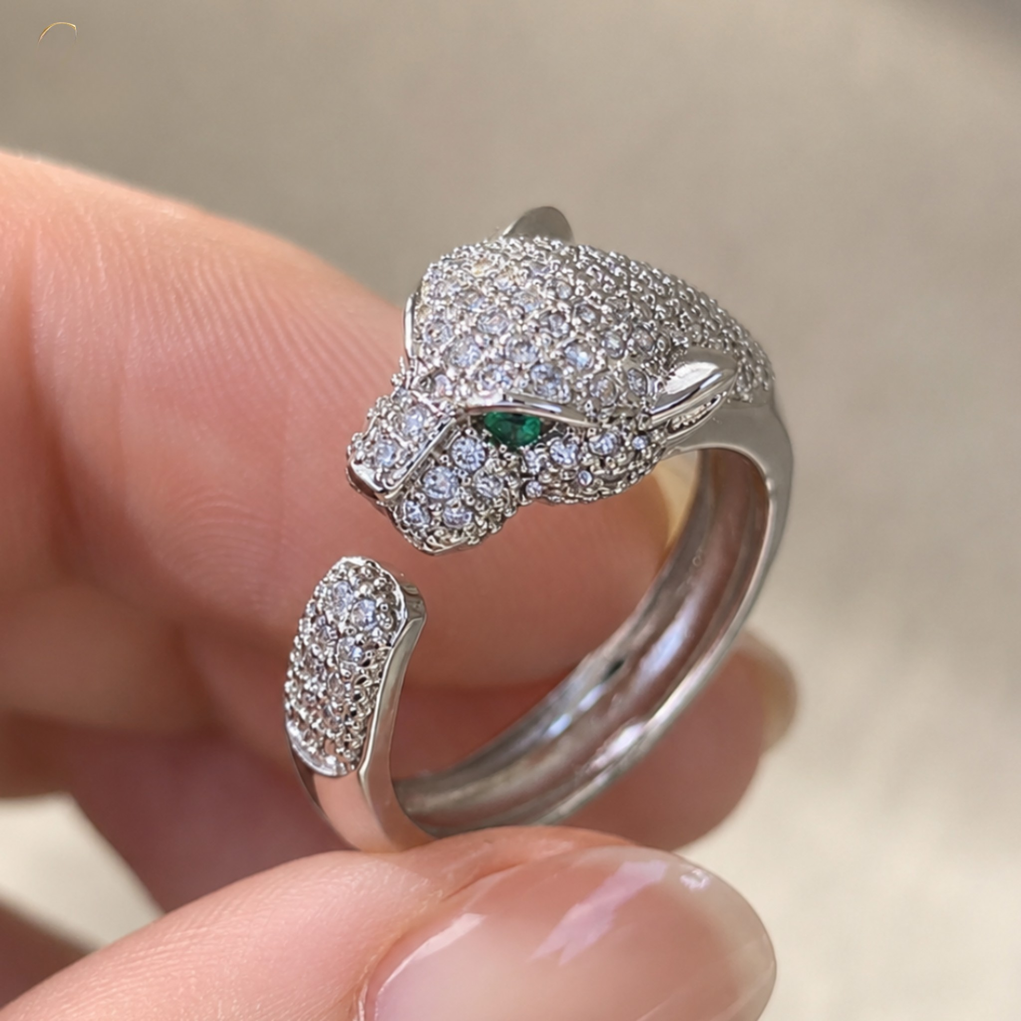 A close-up of a hand holding a silver open ring designed like a panther head, heavily set with clear pavé stones and featuring a small green emerald accent in one eye.