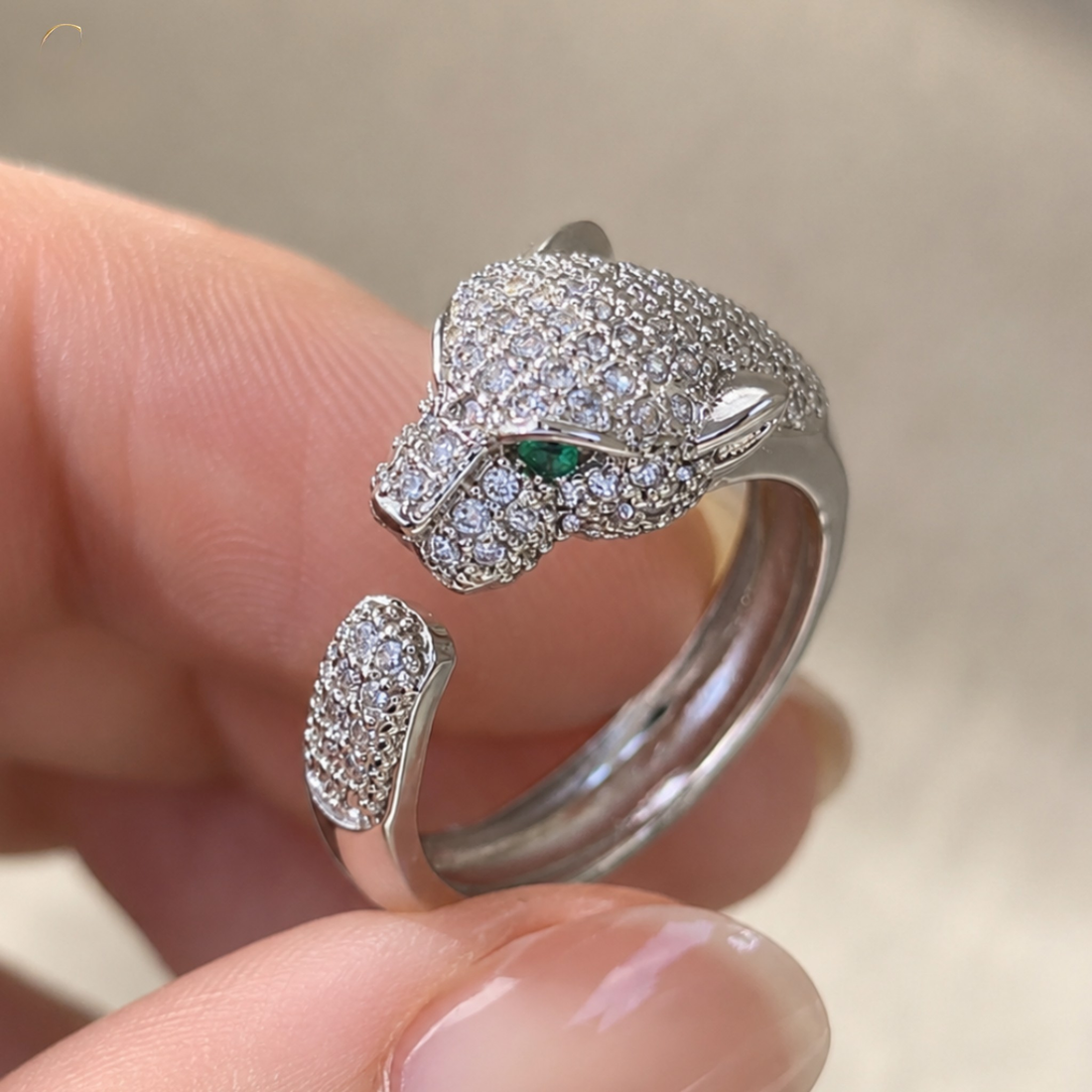 A close-up of a hand holding a silver open ring designed like a panther head, heavily set with clear pavé stones and featuring a small green emerald accent in one eye.