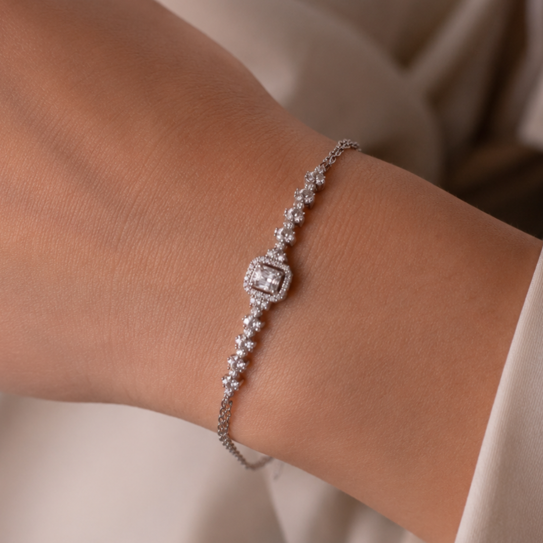 A close-up of a delicate silver-toned bracelet with clear gemstones worn on a person's wrist, set against a soft beige background.