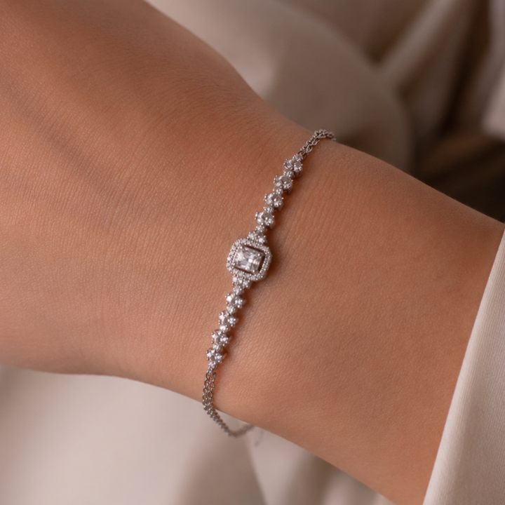 A close-up of a delicate silver-toned bracelet with clear gemstones worn on a person's wrist, set against a soft beige background.