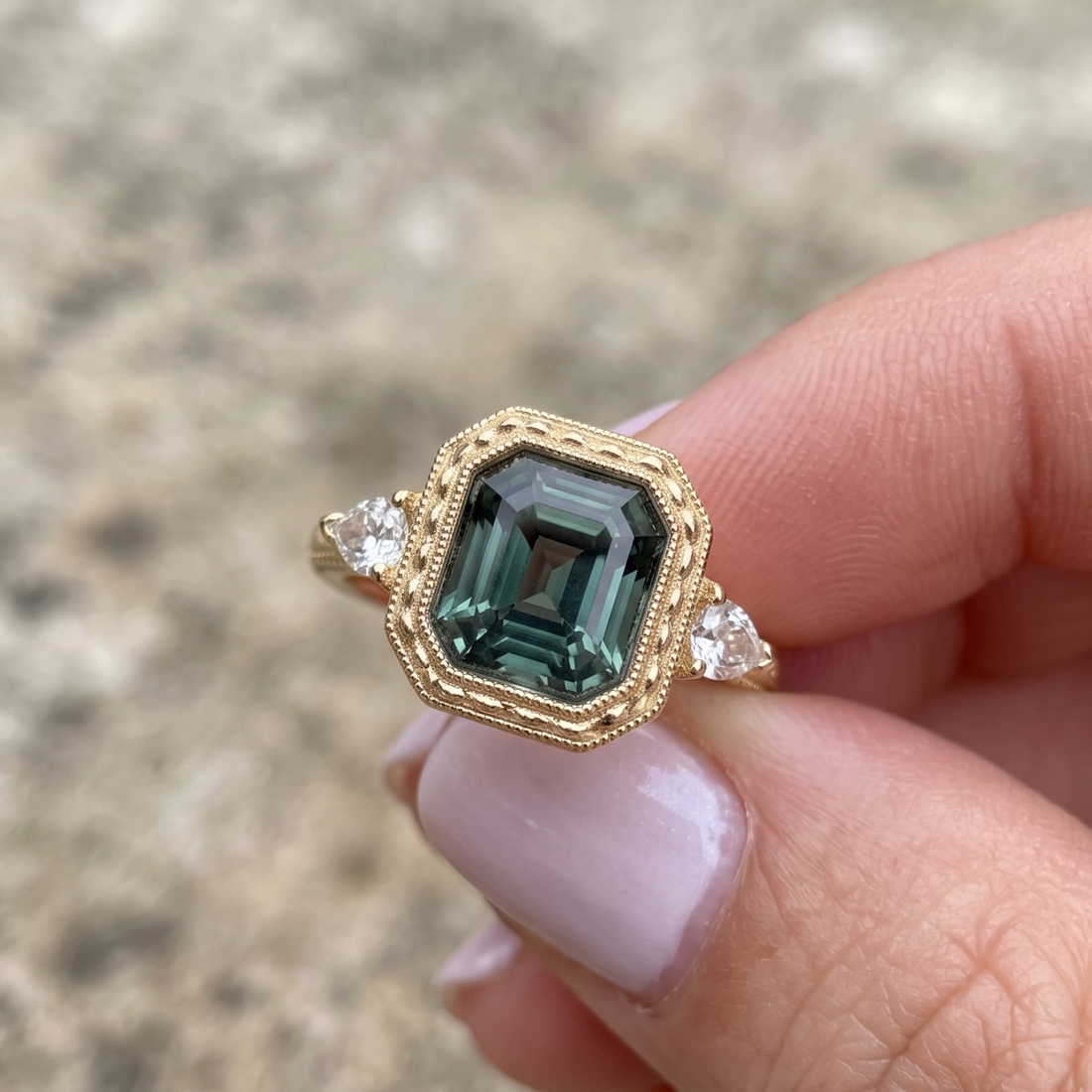 A gold ring with a large emerald-cut green gemstone and two small clear side stones, held between fingers against a blurred outdoor background.