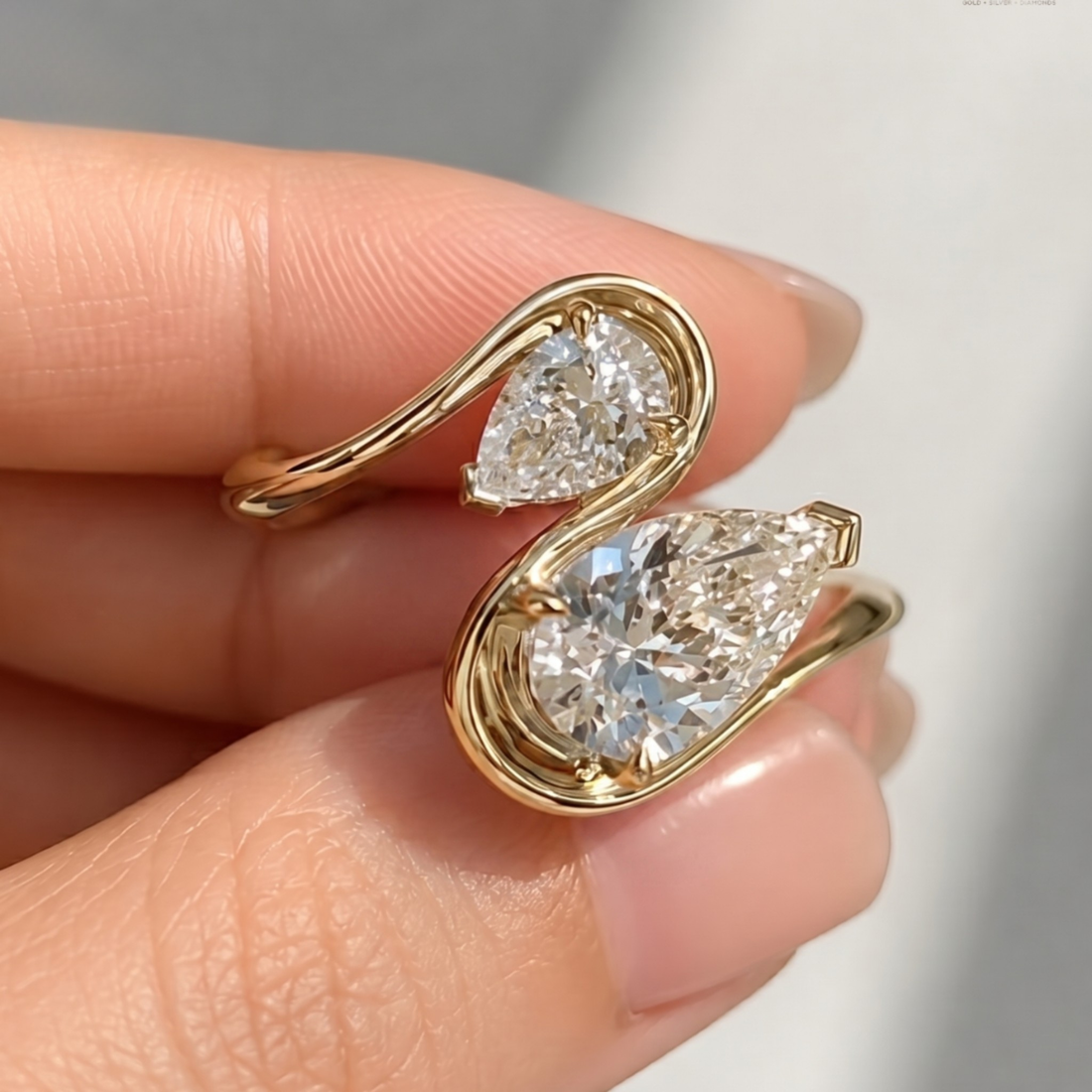 A close-up of a hand holding a gold ring with two pear-cut clear gemstones in a swan-like curved setting.