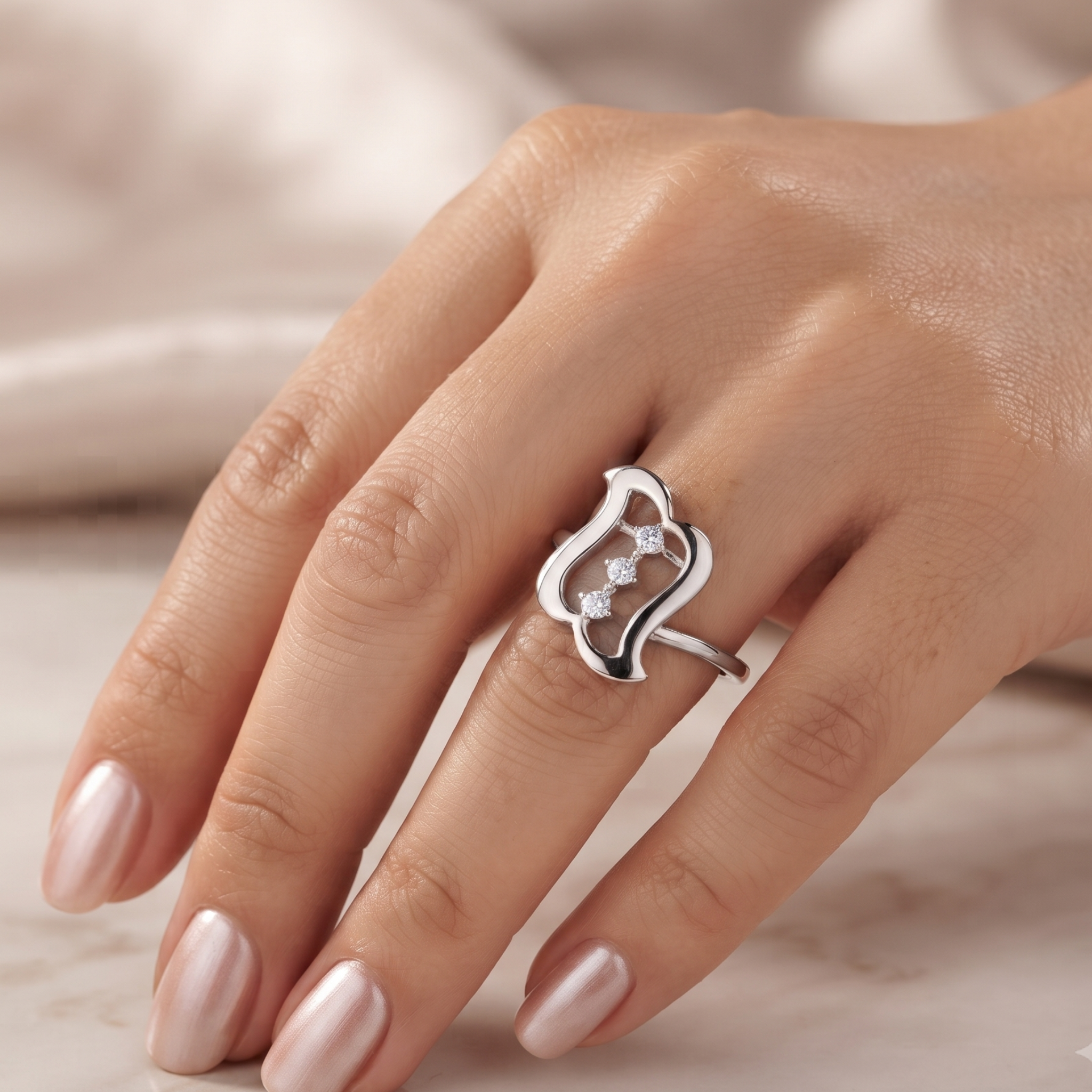 A silver ring with an openwork abstract design and small clear stones, shown worn on a hand with pale pink manicured nails against a soft beige background.